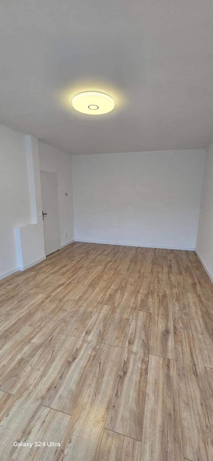 Lege kamer met witte muren, houten vloer en plafondlamp.