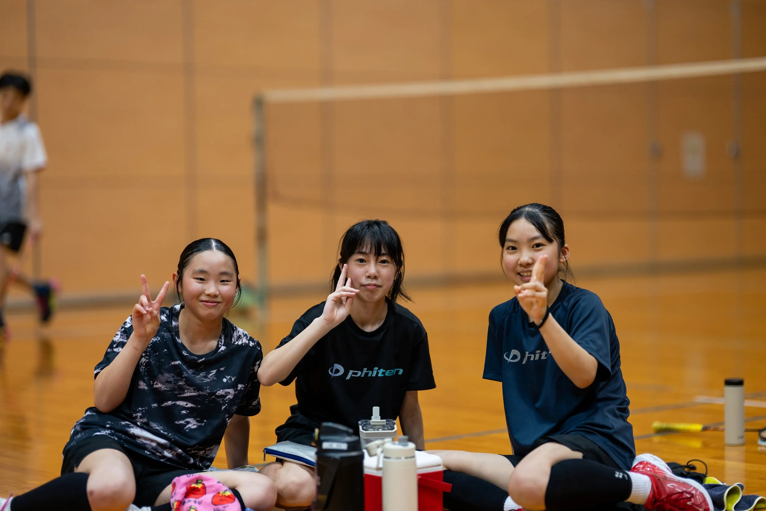 バドミントンの室内コートで座っている3人の若い女性選手が笑顔でポーズをしている。山形県バドミントンクラブチーム、小学生、中学生の部活動地域移行鶴岡市