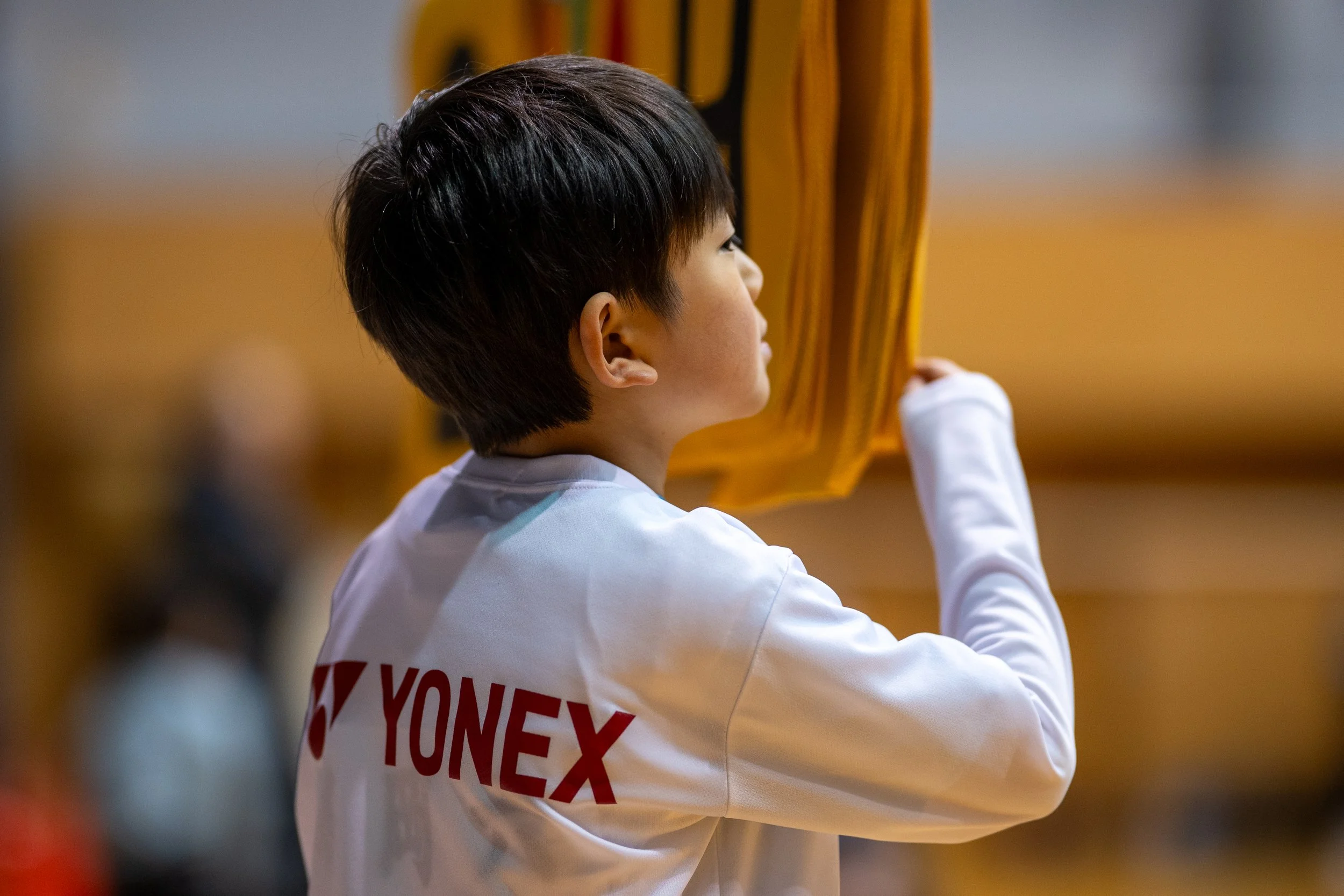 白いスポーツジャケットの少年が、背中に赤字のロゴYONEXと共に、黄色のバドミントンラケットを持っている。