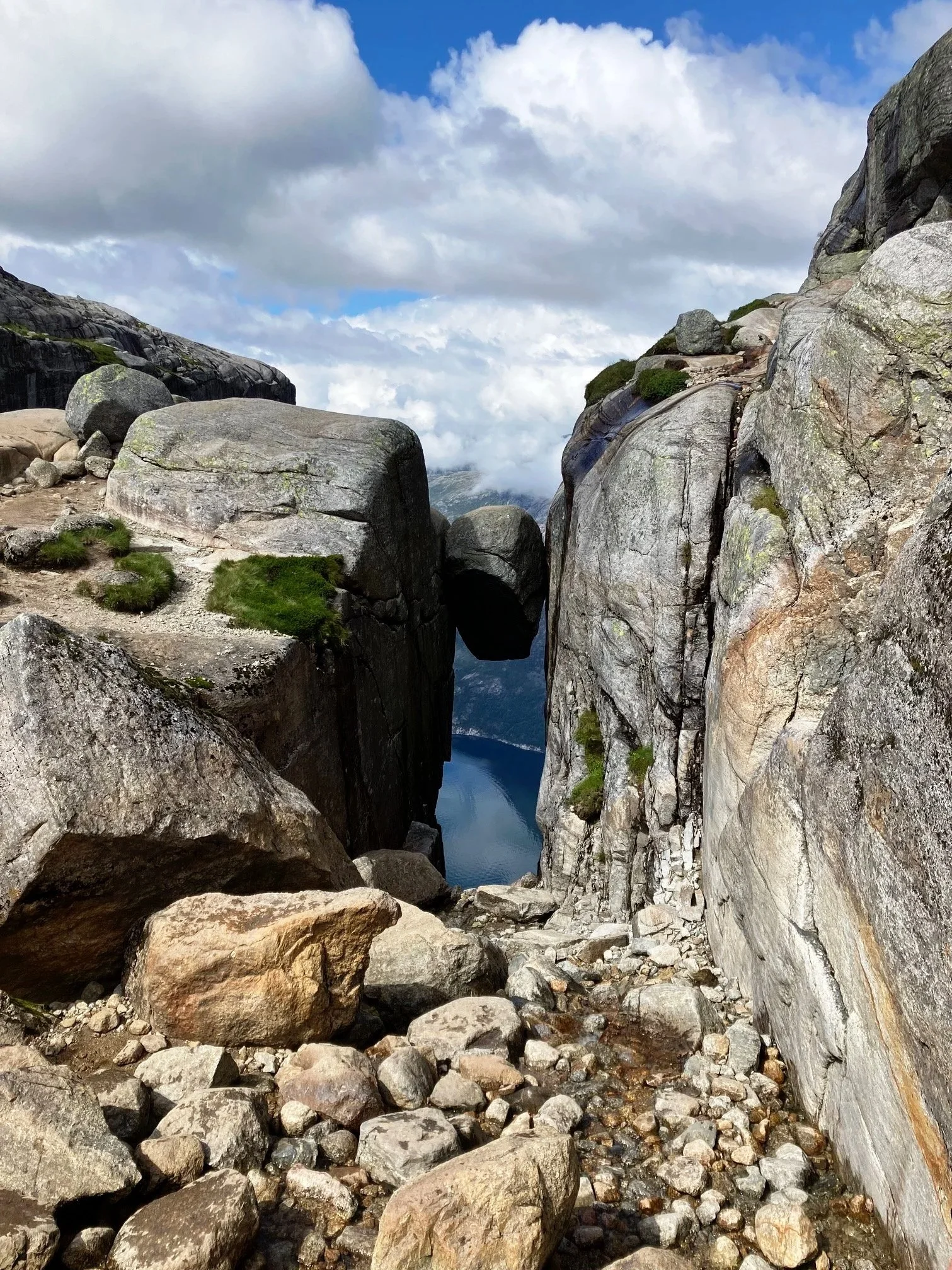 A narrow rocky passage between large granite boulders overlooking a fjord or lake surrounded by mountains, under cloudy sky.