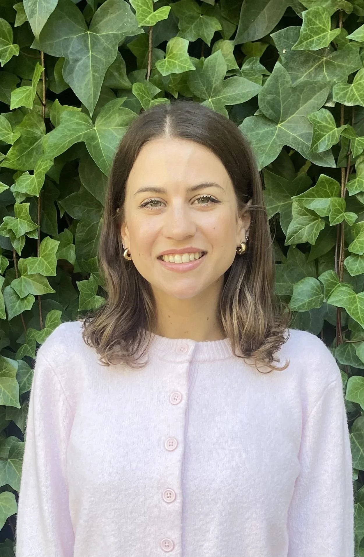 A woman with shoulder-length wavy brown hair, light skin, and hoop earrings smiling, standing in front of a lush green ivy wall.