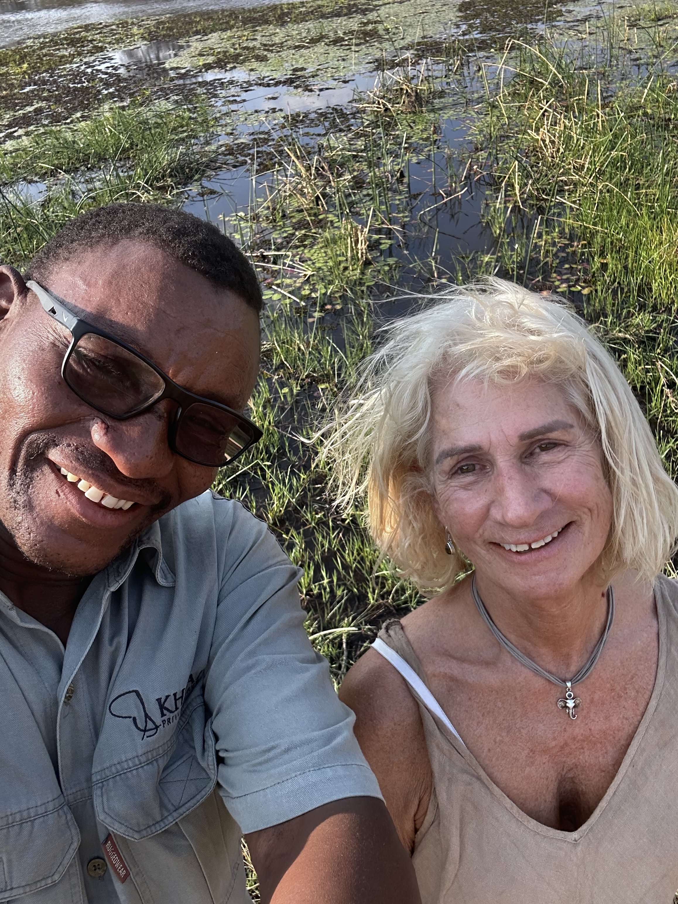 Two smiling people taking a selfie outdoors near a pond with aquatic plants.