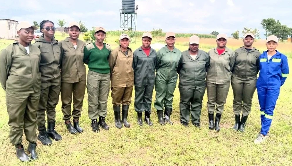 A group of ten women in field uniforms standing outdoors, with some wearing hats and rain boots, and one in a blue safety jumpsuit, on a grassy area with a tower and trees in the background.