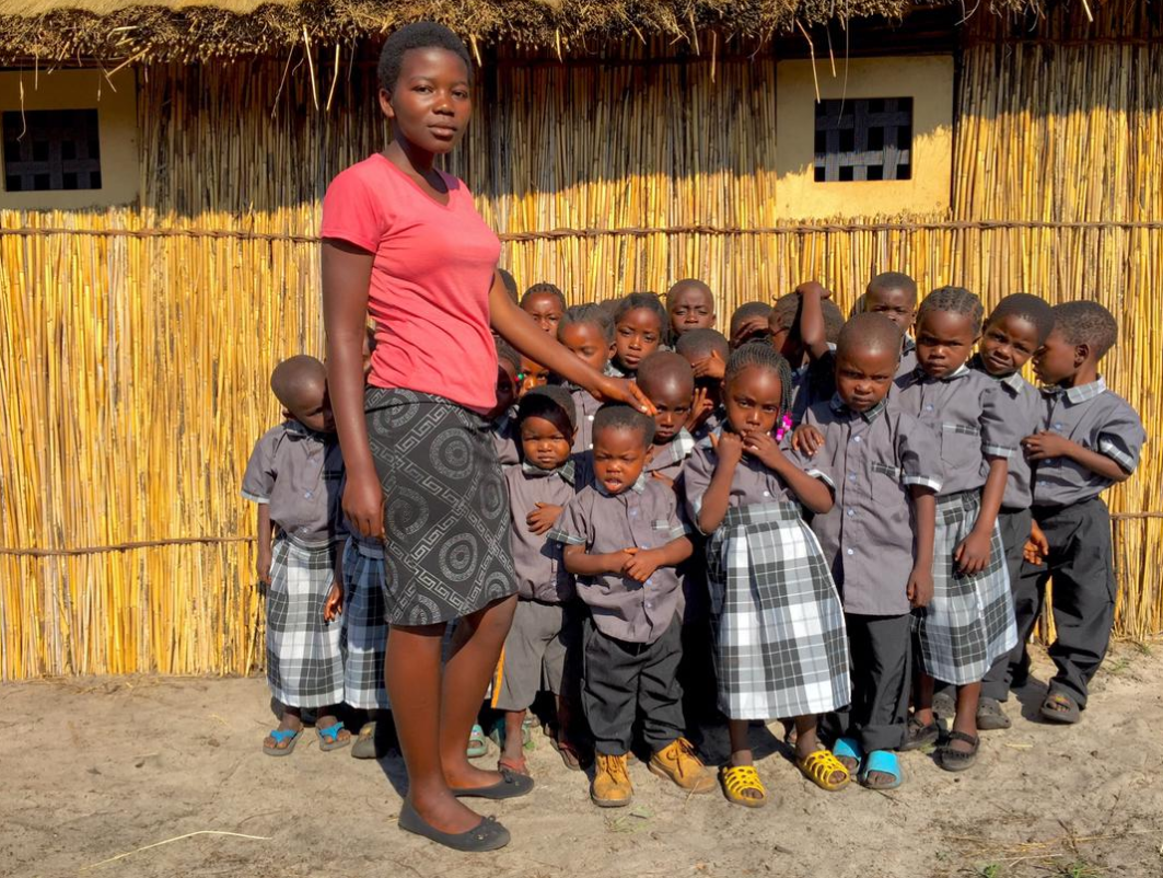 A young woman stands in front of a group of children outside, with a straw fence and a thatched roof building in the background. The children are wearing matching uniforms and are looking at the camera.