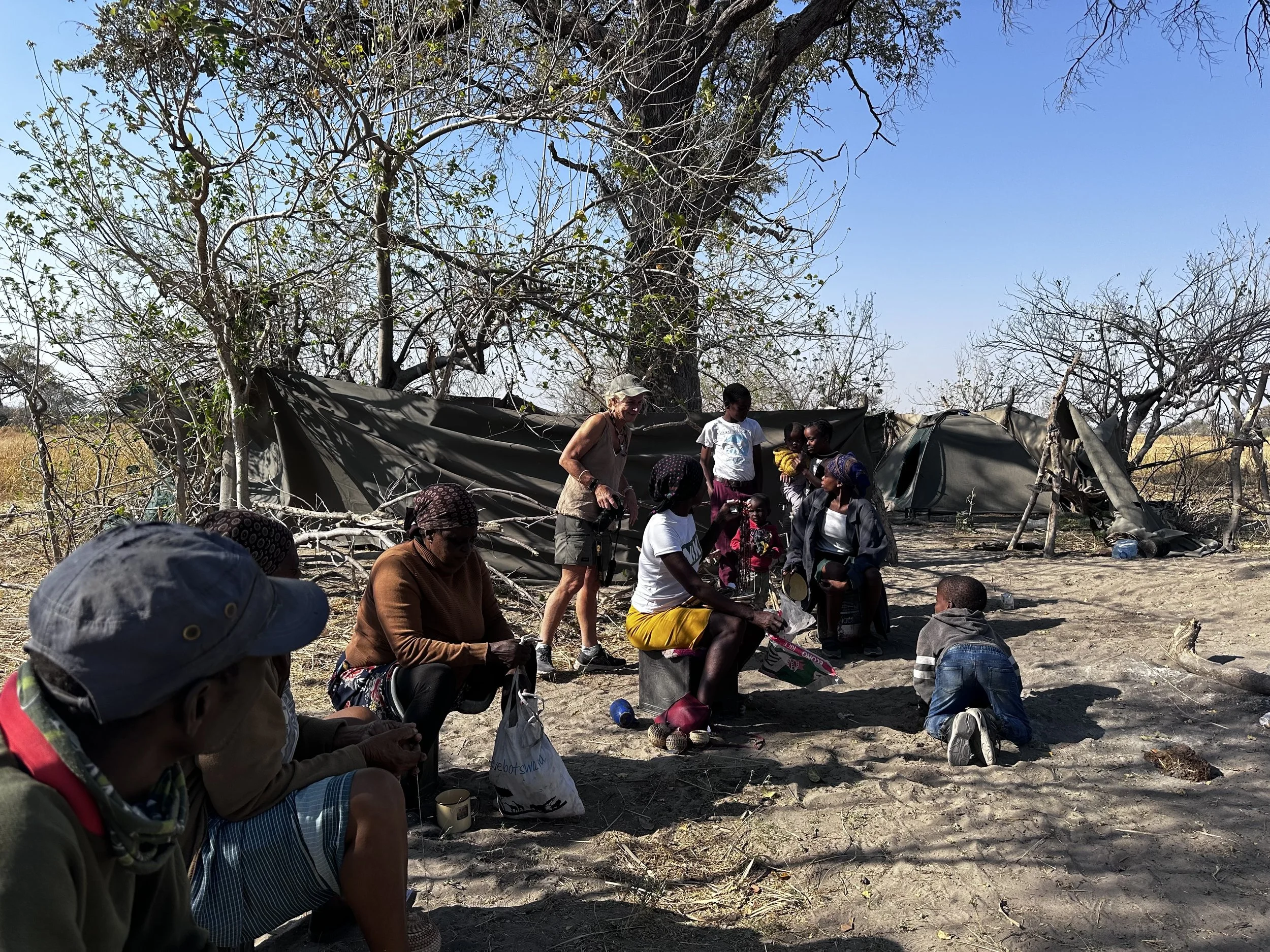 A group of people gathering outdoors under a large tree with few leaves, surrounded by dry grass and scattered branches, some sitting and some standing, with makeshift tents in the background on a sunny day.