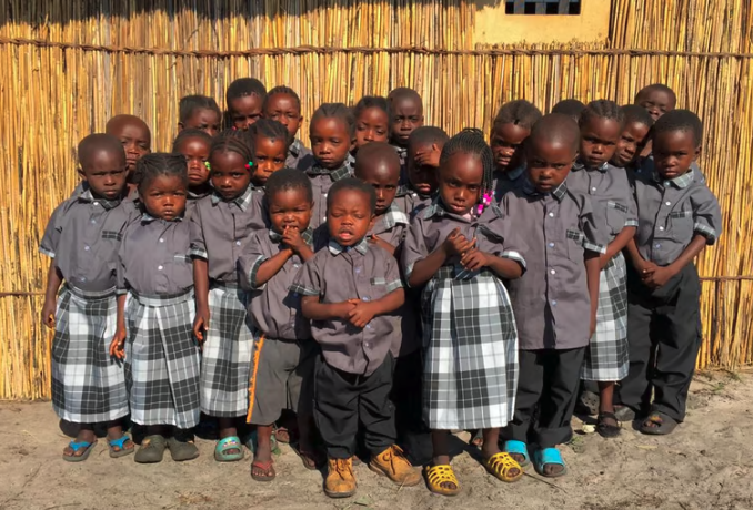Group of young children in school uniforms standing outdoors in front of a bamboo fence.
