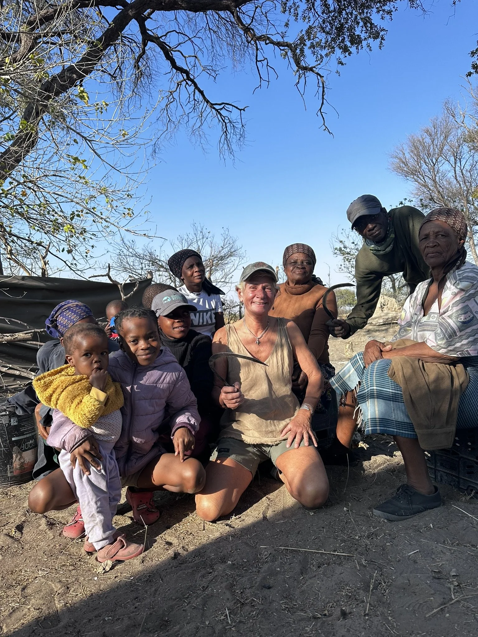 A group of people, including children and adults, gathered outdoors on a dirt ground with leafless trees in the background. The scene appears to be a community gathering or a meeting.