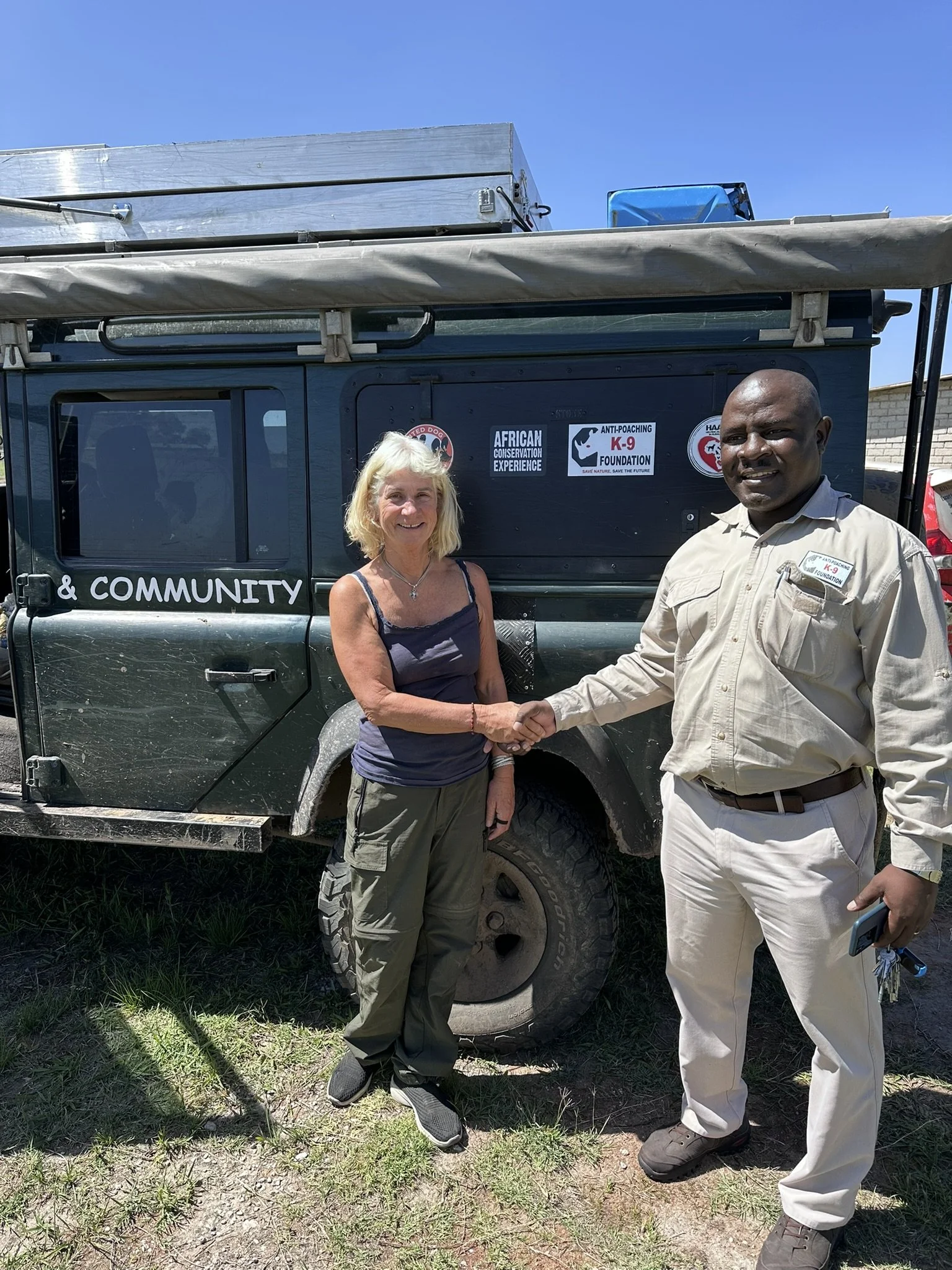 A woman and a man are shaking hands in front of a black off-road vehicle with various logos, including 'AFRICAN CONSERVATION EXPERIENCE' and 'ANTI-POACHING K-9 FOUNDATION'. They are outdoors on a sunny day.