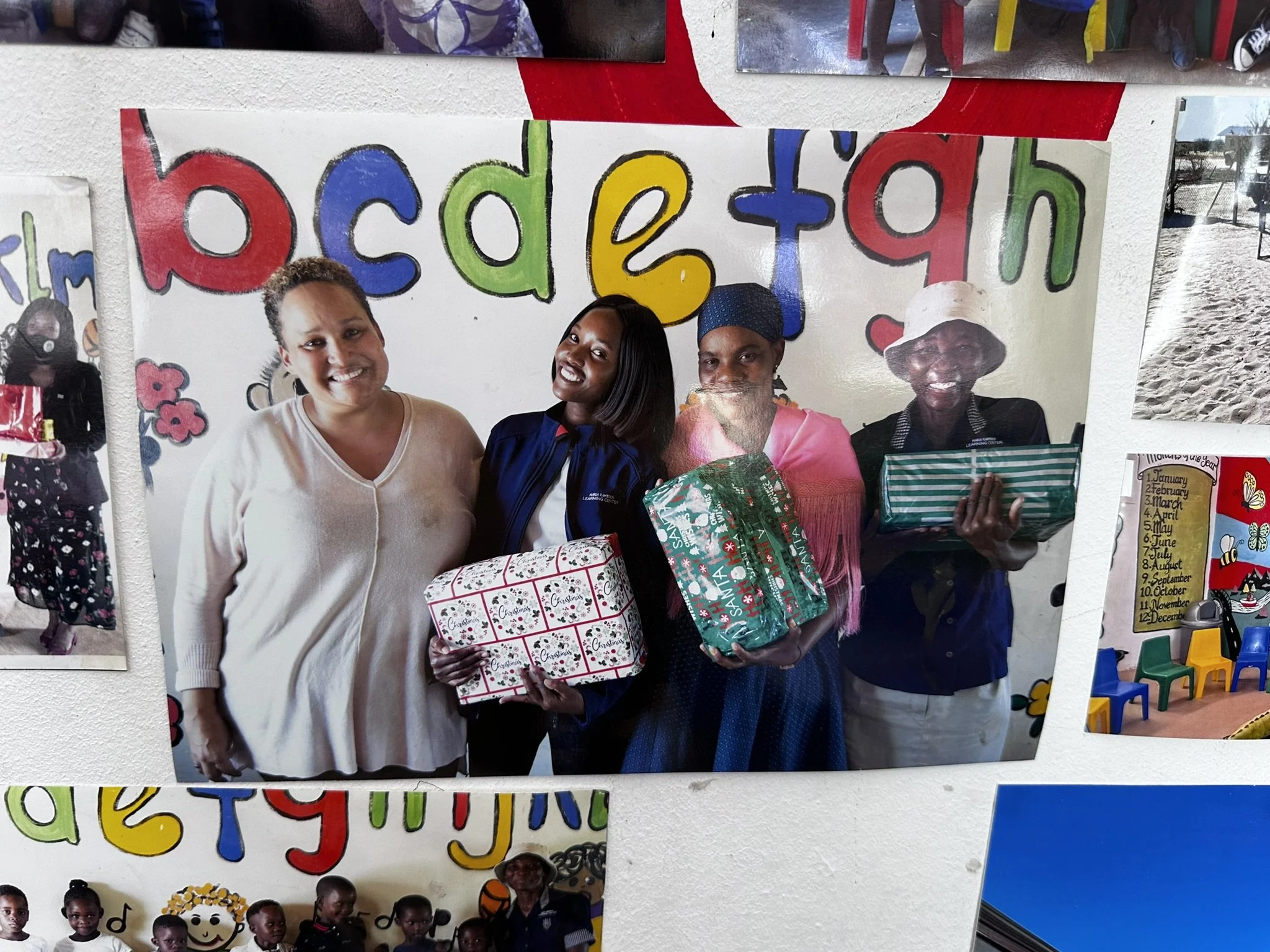Photo of four women smiling, holding wrapped Christmas gifts, standing in front of a colorful painted wall that says "education" and features children at the bottom.