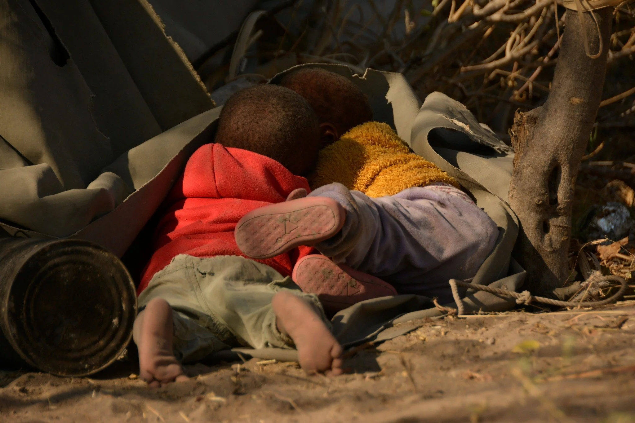 A person lying on the ground, covered with a yellow blanket, with two children leaning on them. The scene appears to be outdoors, with tree branches around and the ground visible. The image is candid and suggests a moment of rest or concealment.
