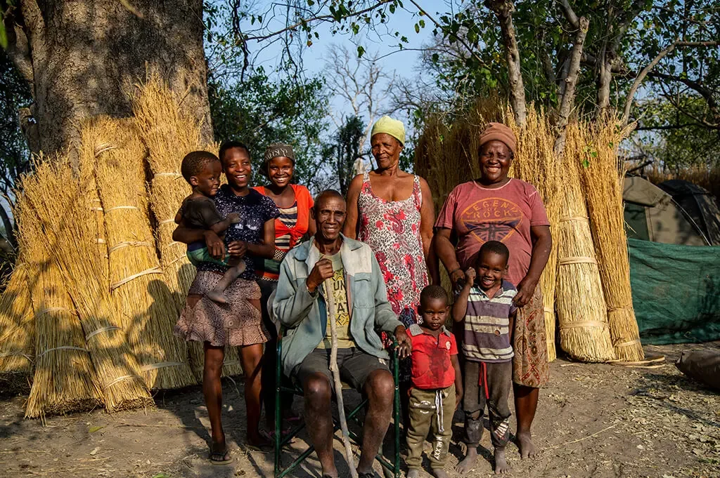 A group of six children and two elderly women outdoors, standing and sitting in front of bundles of harvested crops, with trees and a tent in the background.