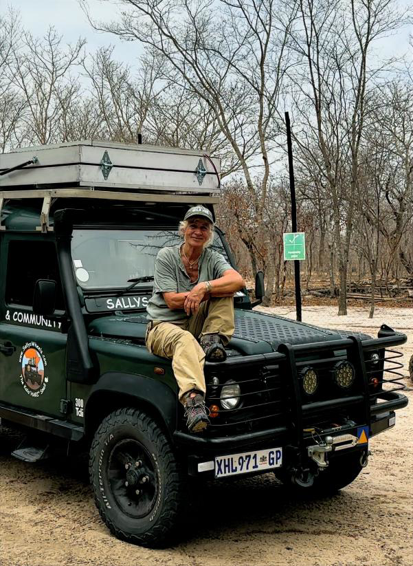 A woman sitting on the front of a dark green safari vehicle in a wooded area. She is smiling and wearing a gray shirt, khaki pants, and a cap. The vehicle has a roof rack with luggage and has an emblem on the side.