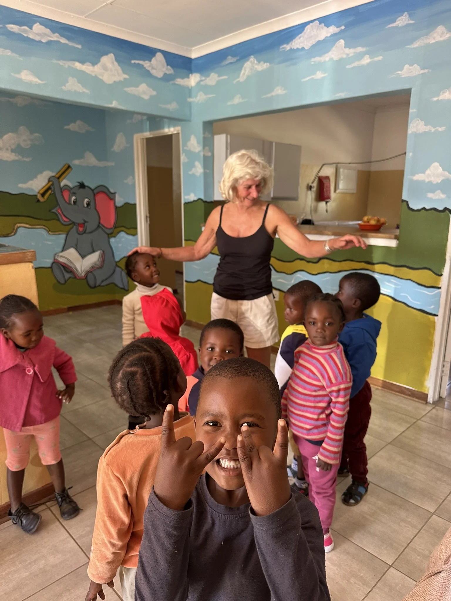 Group of children with a woman in a colorful room with a mural of an elephant holding a pencil and reading a book. The kids are smiling and engaging with the camera.