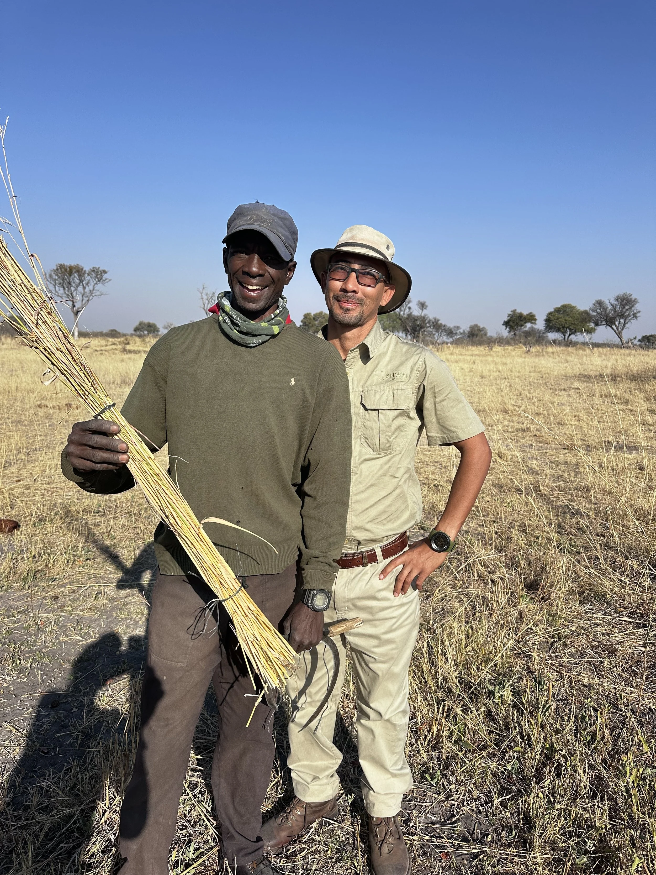 Two men standing in a grassy, open field under a clear blue sky, one holding a bunch of harvested grass or reeds, both smiling and dressed in outdoor clothing with hats.