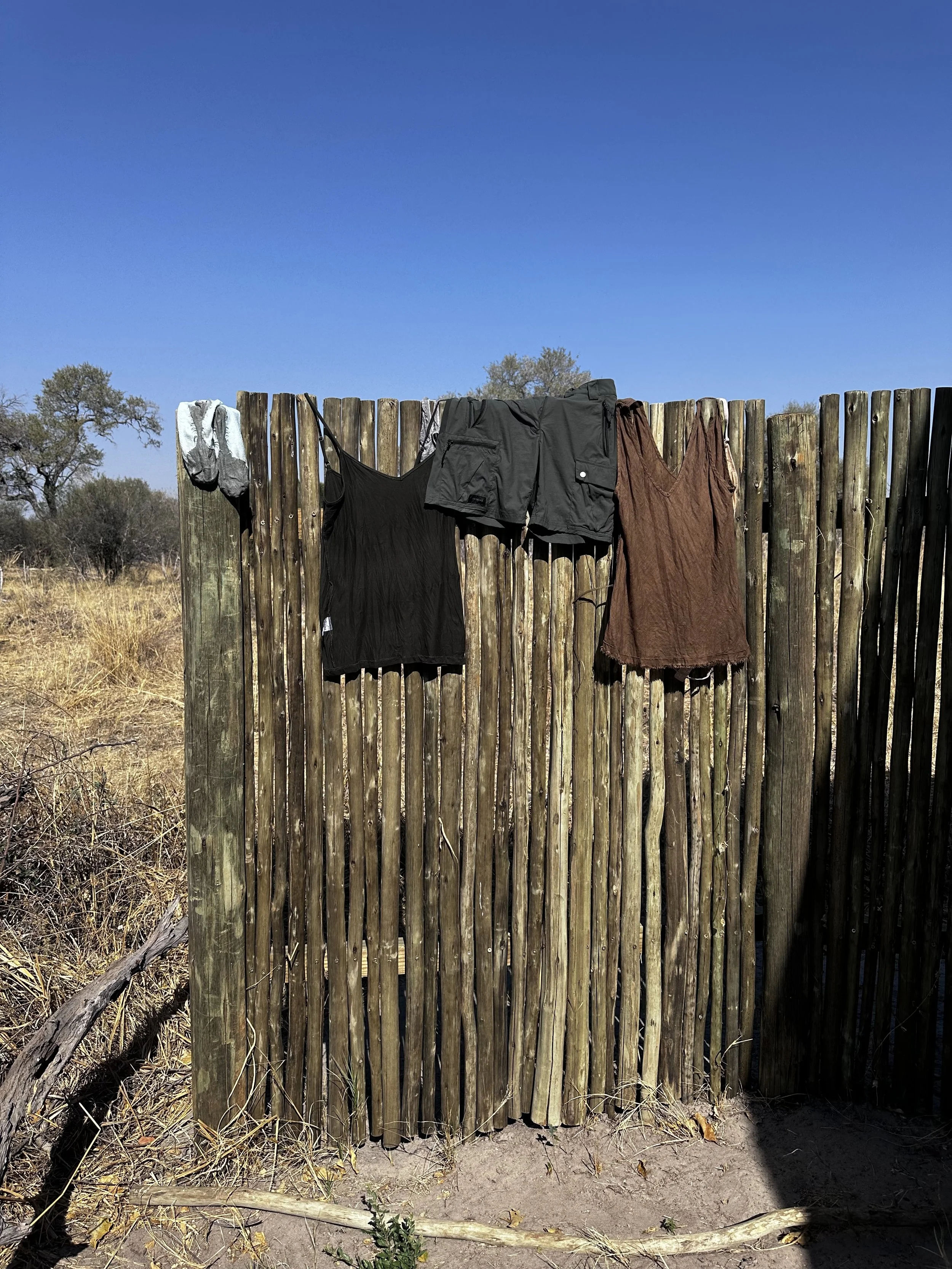 Clothes hanging on a wooden fence, including a pair of grey socks, a black top, black shorts, and a brown dress, with a clear blue sky and trees in the background.