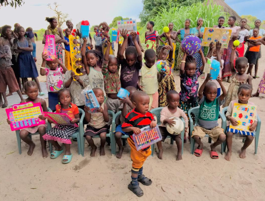 A group of children and adults outdoors, with children holding colorful educational toys, some sitting in chairs and others standing in the background.