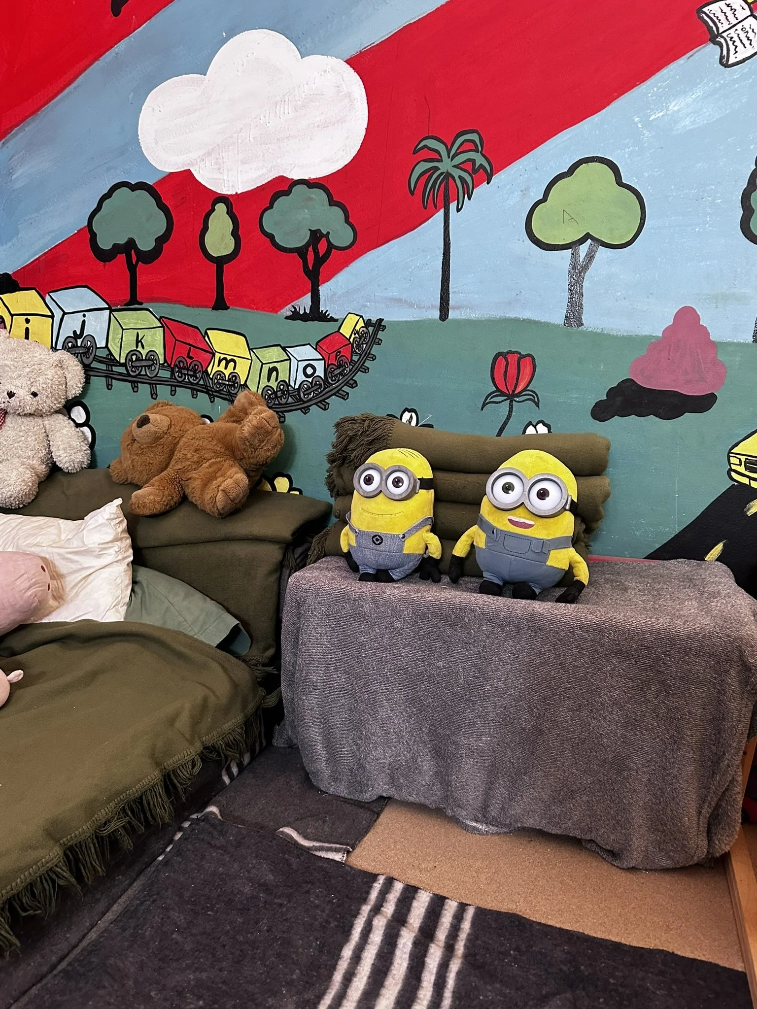 A colorful mural of trees, clouds, flowers, and a train with stuffed animals and plush toys in front of a couch and a blanket.
