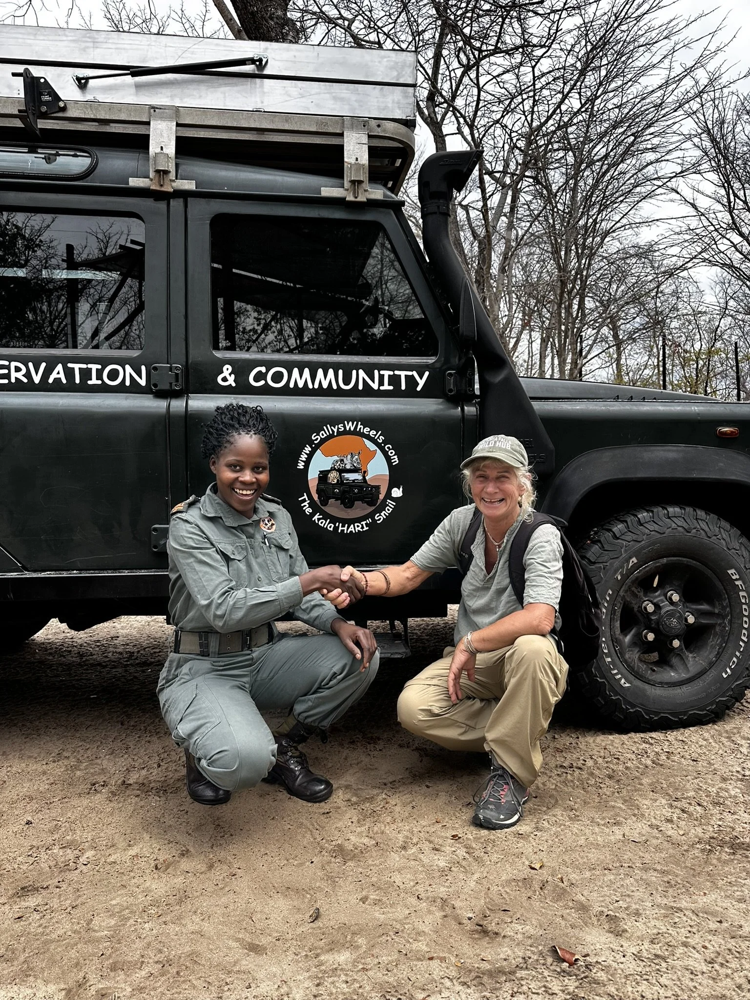 Two women, one in a gray ranger uniform and the other in a tan outfit, are smiling and shaking hands in front of a black off-road vehicle with a logo of a leopard on the door. The vehicle has a roof rack and a snorkel and is parked on dirt, with leafless trees in the background.