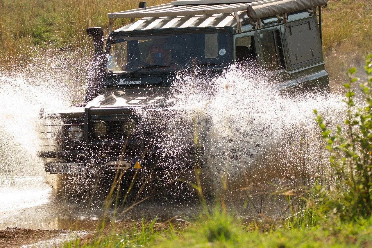A black off-road vehicle driving through a muddy water puddle, splashing water as it moves.