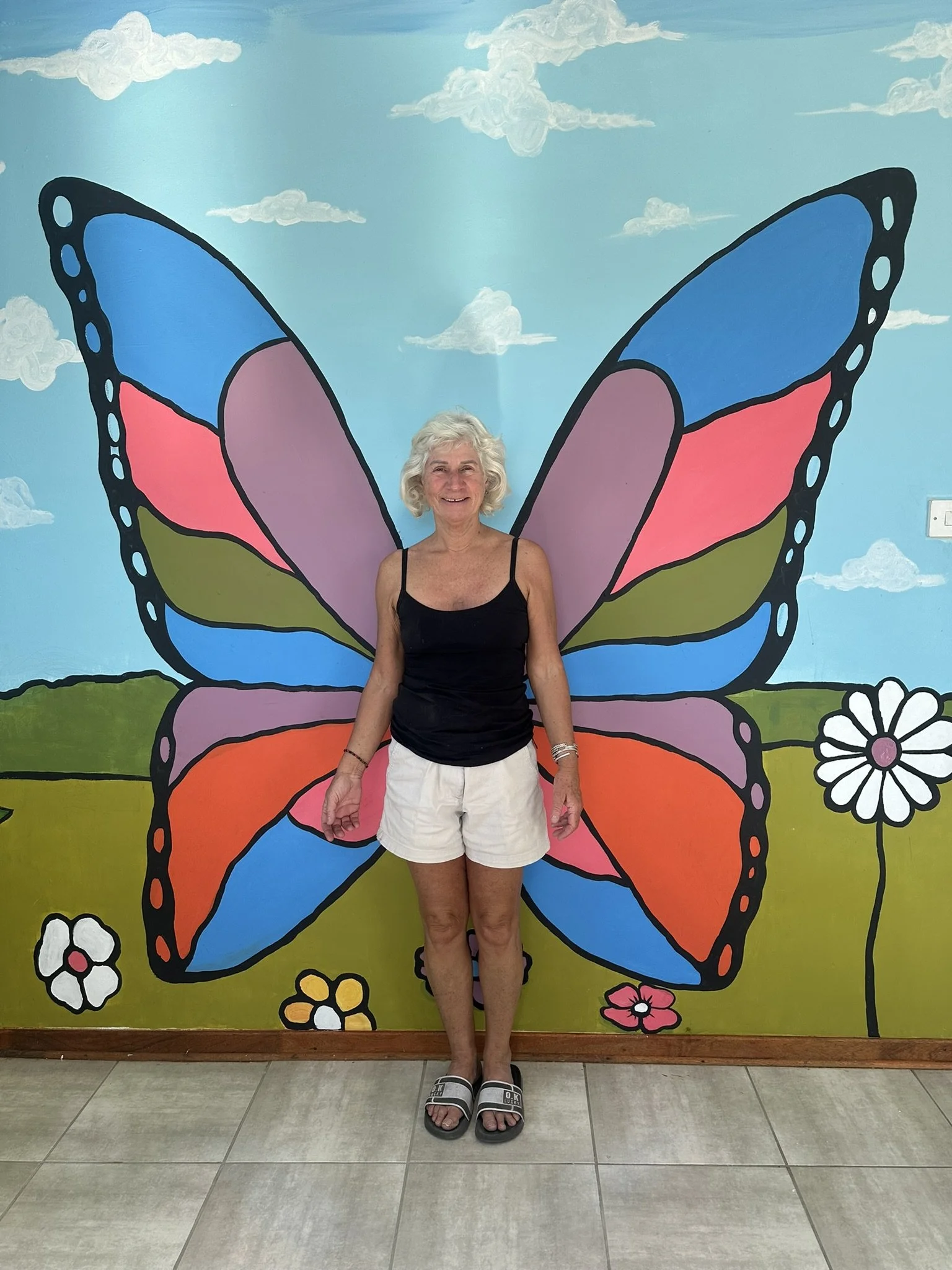 A smiling elderly woman with curly white hair standing in front of a colorful butterfly mural on the wall. The woman is wearing a black tank top, white shorts, and black sandals, and is indoors on a tiled floor.