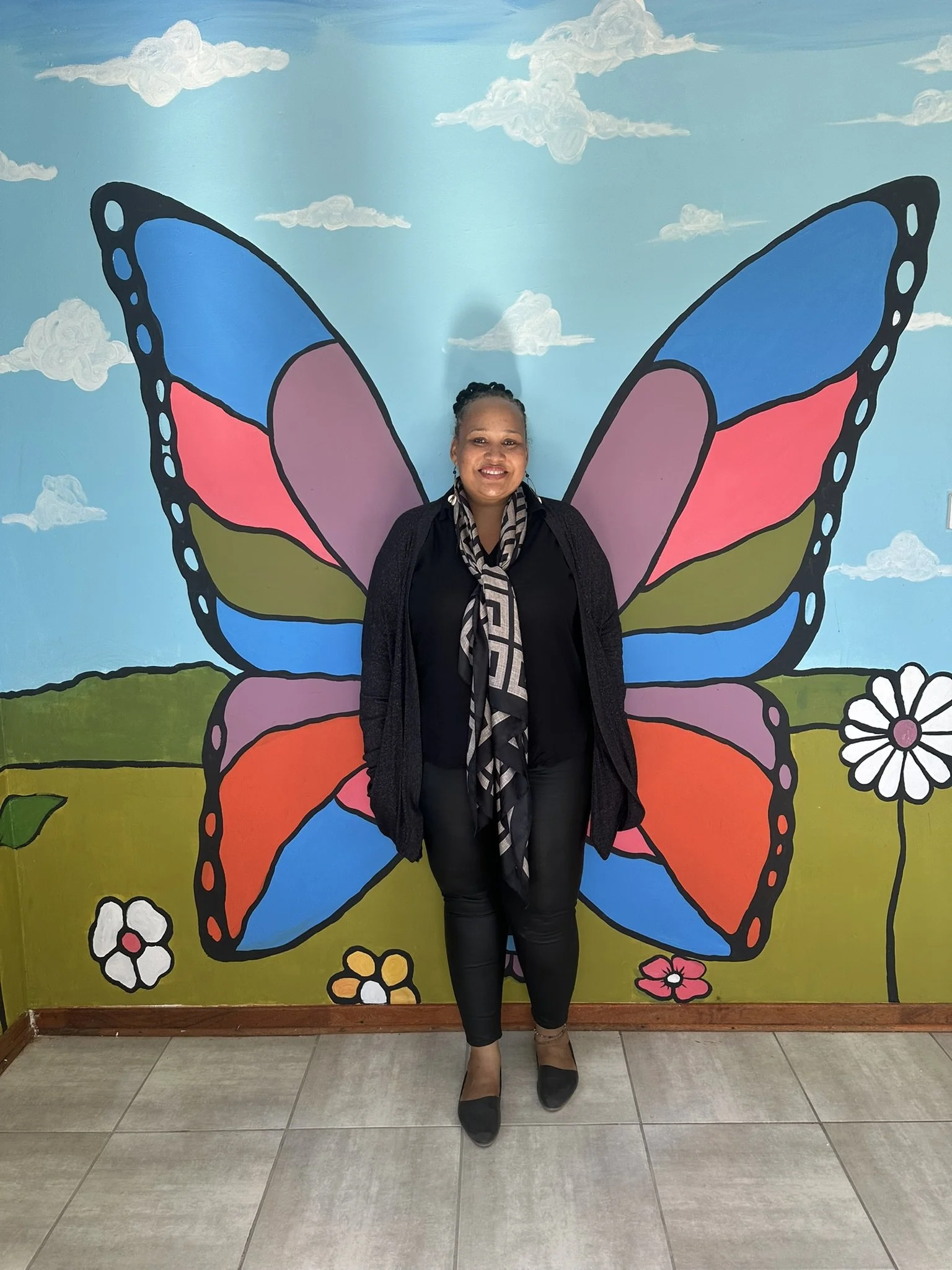 A woman standing in front of a colorful butterfly mural on a wall, with flowers and clouds painted around it.