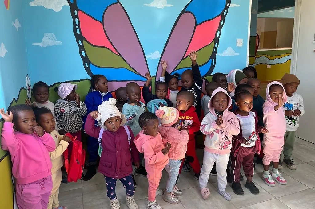 A group of young children standing in front of a colorful wall with a large butterfly mural, some children are raising their hands or making gestures.