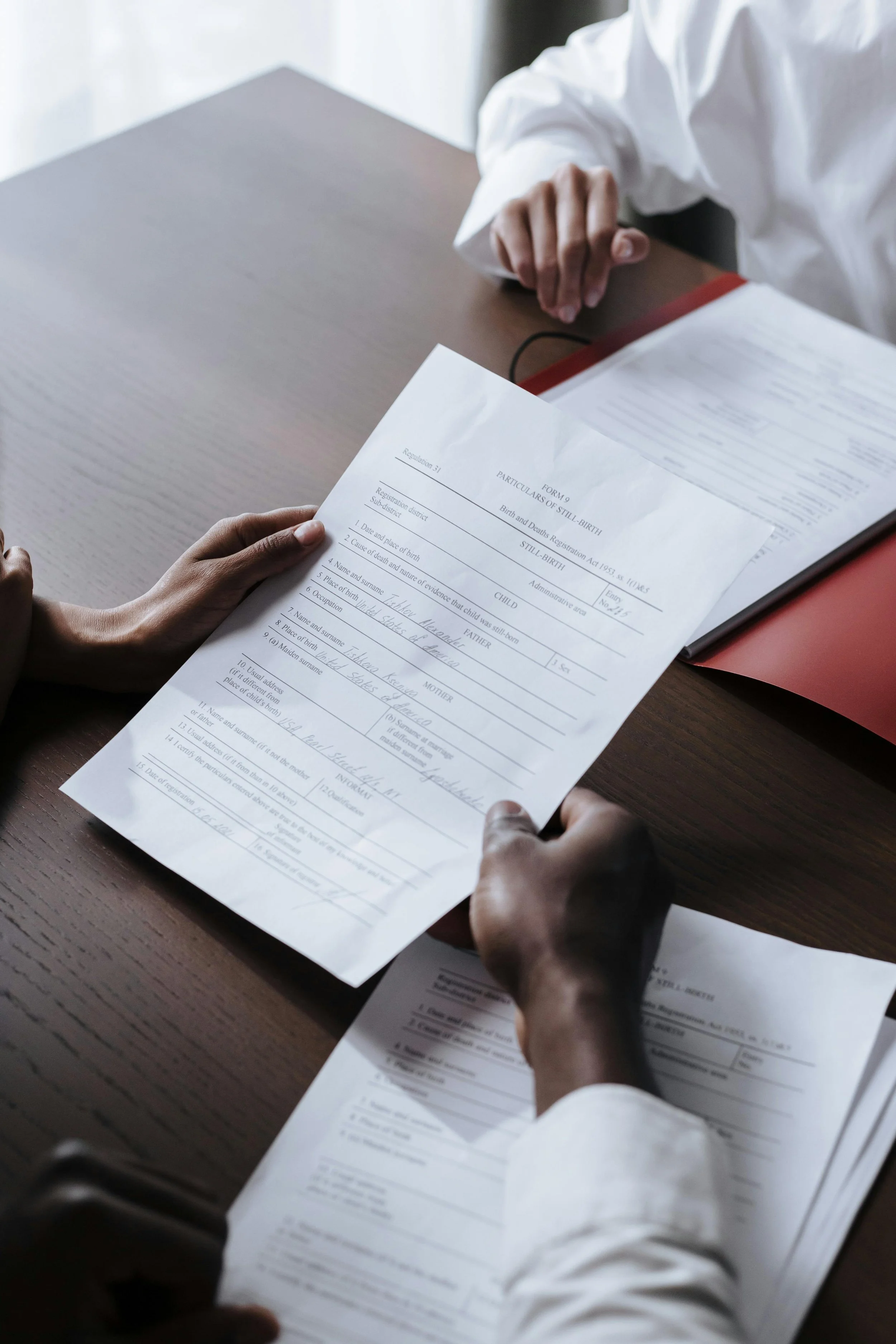 Two people sitting at a wooden table, holding and reviewing a birth registration form with handwritten entries.