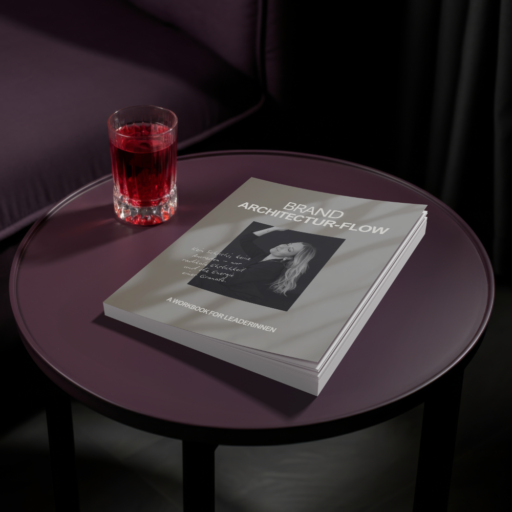 Ein Hellgraues Buch mit dem Titel 'BRAND ARCHITECTUR-FLOW' liegt auf einem samtigen, lila Tisch. Auf dem Buch ist ein schwarz-weiß Bild einer Frau zu sehen. Neben dem Buch steht ein Glas mit rotem Getränk. Im Hintergrund sind dunkle Vorhänge und ein Sitz.