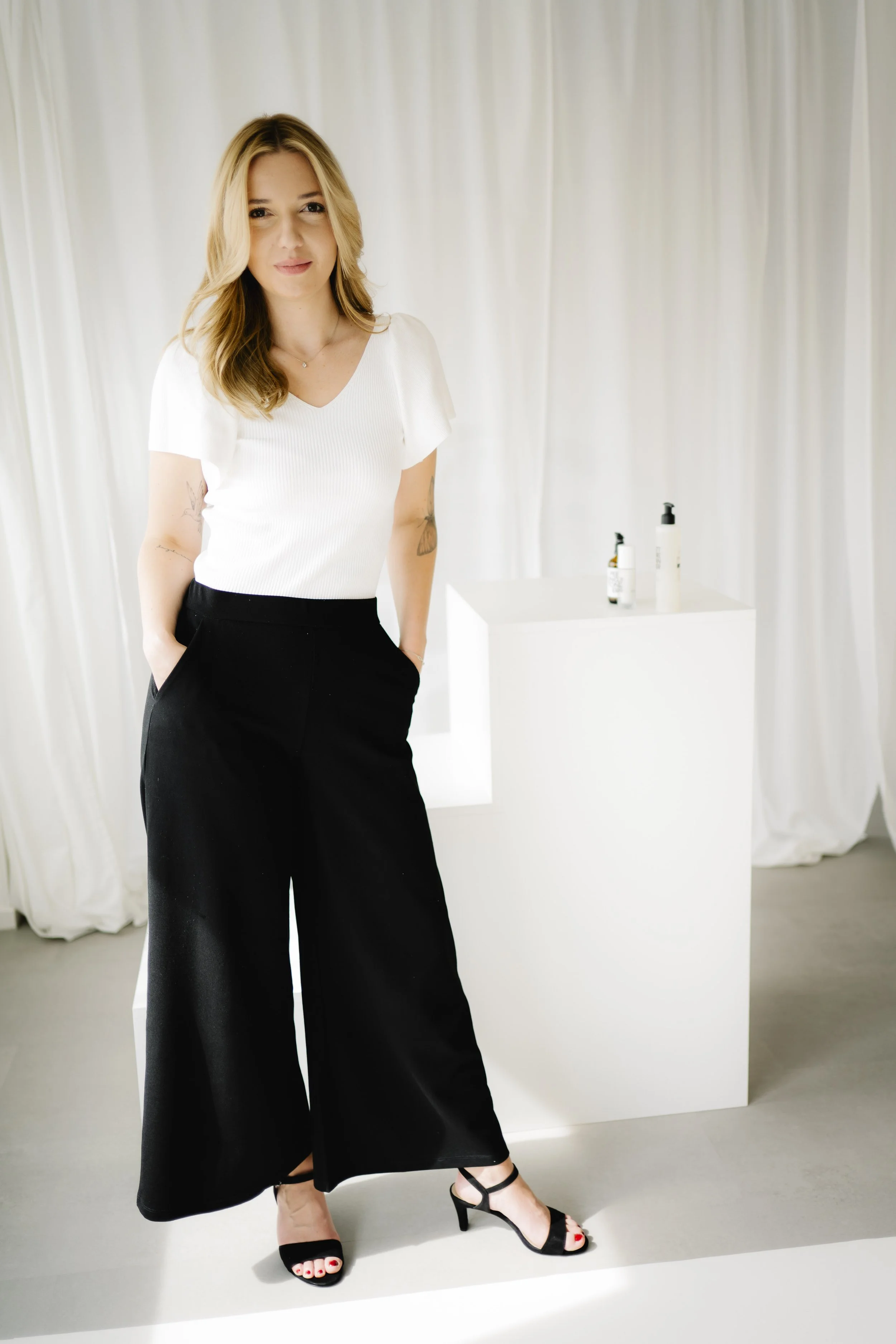 Eine Frau mit blonden Haaren in einem weißen Oberteil und schwarzen weiten Hosen, die auf hohe Schuhe steht, vor weißen Vorhängen und einem weißen Podest mit Hautpflegeprodukten.