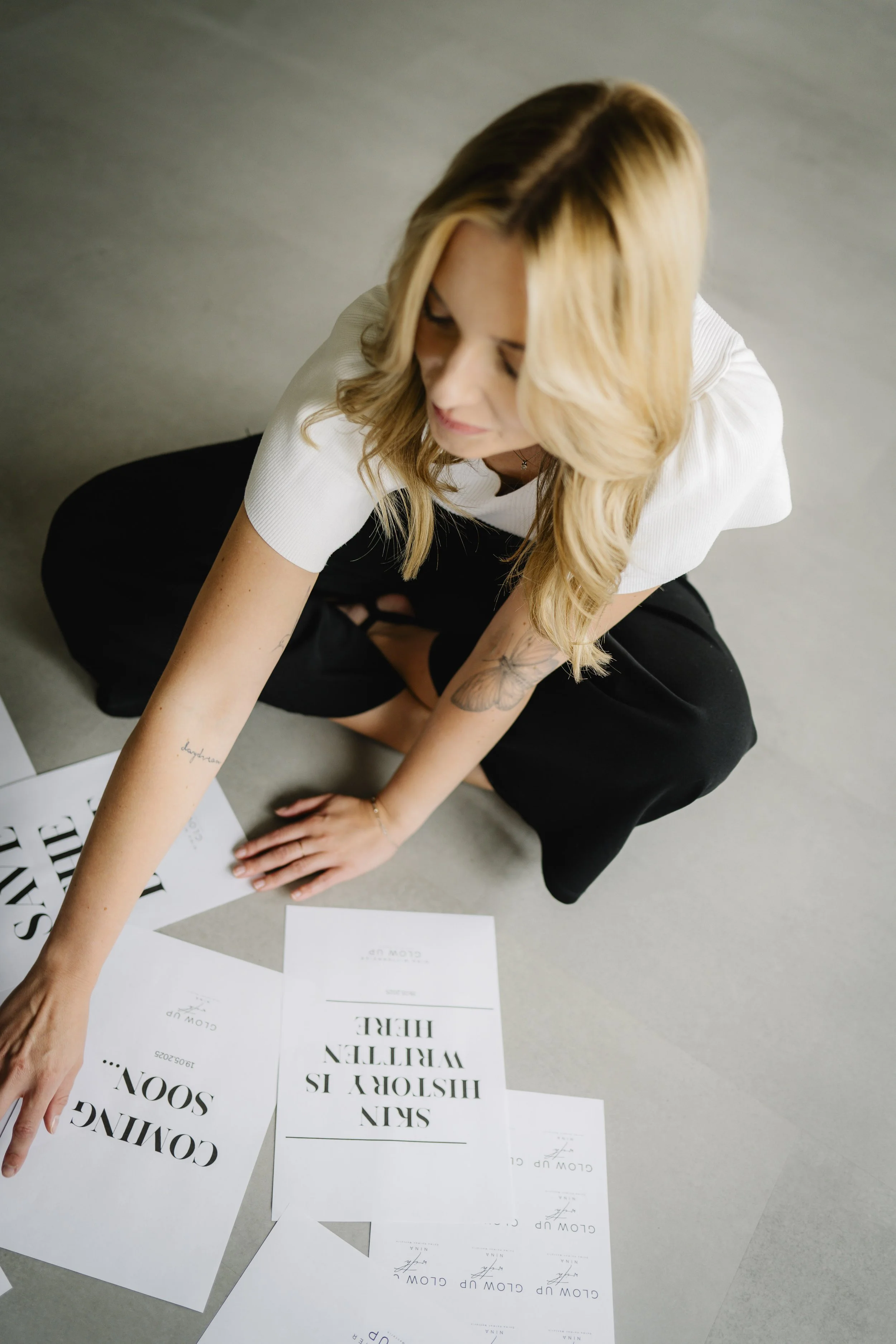 Eine blonde Frau sitzt auf dem Boden, schaut nach unten und ist umgeben von weißen Papierbappen mit Texten wie "CON GANG SOON..." und "SAY ISHITEN WELCOME HERE".