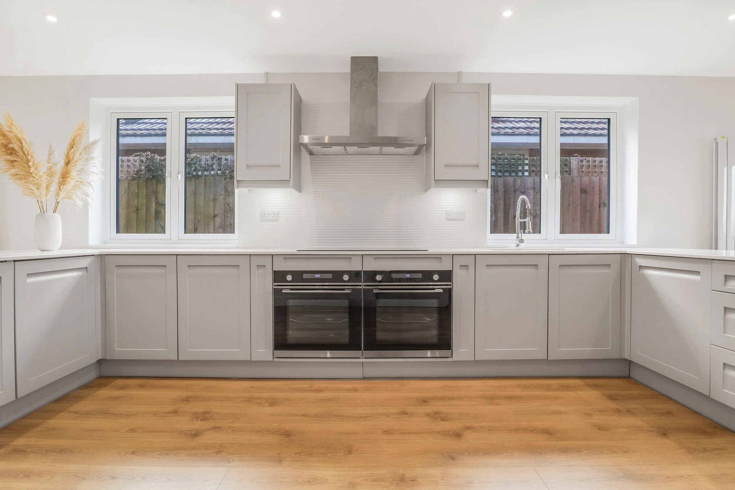Modern kitchen with gray cabinets, a metallic range hood, and two ovens, with two windows showing an outdoor fence and sky.