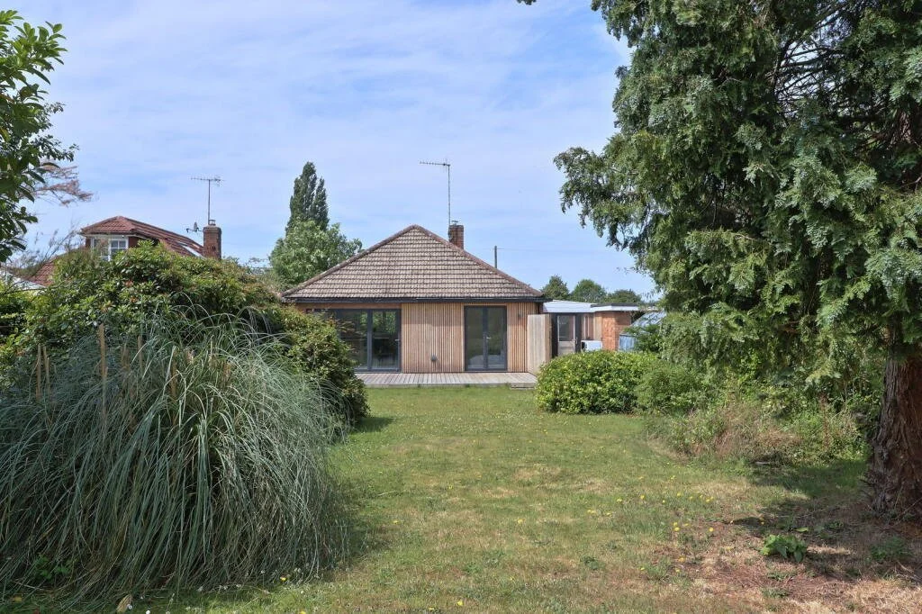 A house with a tiled roof and large glass sliding doors, surrounded by trees, shrubs, and a grassy lawn.