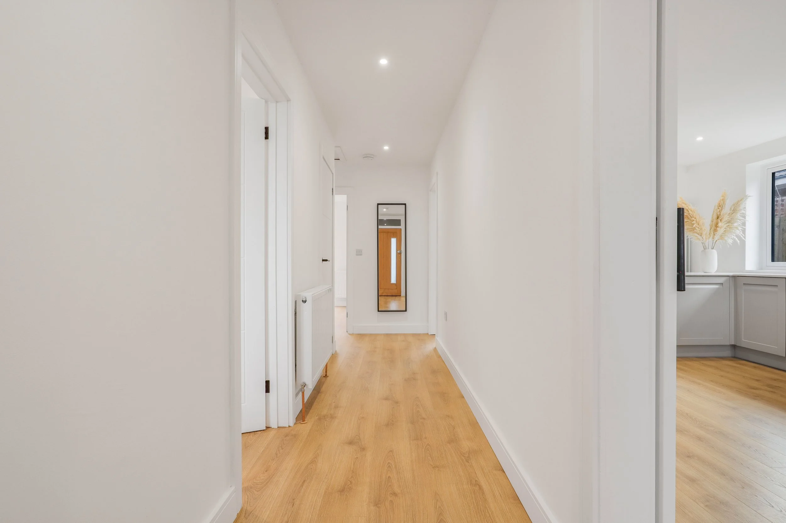 Empty hallway with white walls, wooden floor, and a full-length mirror on the wall at the end, leading to a door with a glass panel.