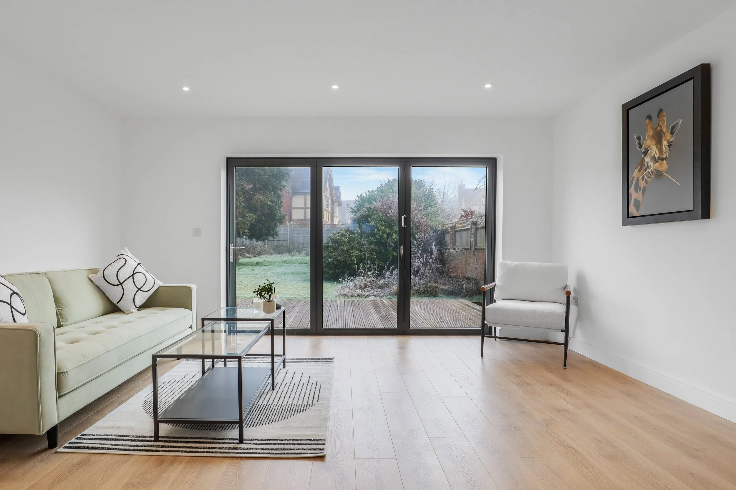 Living room with a glass sliding door leading to a garden, white walls, light-colored hardwood floor, a pale green sofa with patterned pillows, a white armchair, a black framed giraffe picture on the wall, a striped rug, and a small coffee table.