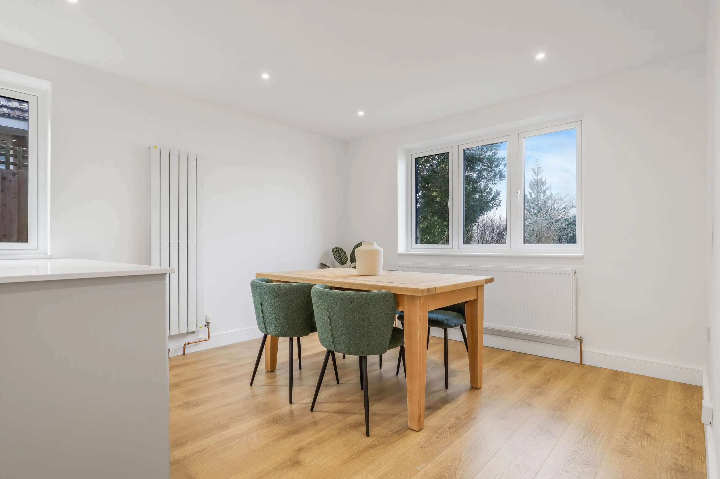 Empty dining room with wooden table, four green chairs, hardwood floors, white walls, and large window showing trees outside.
