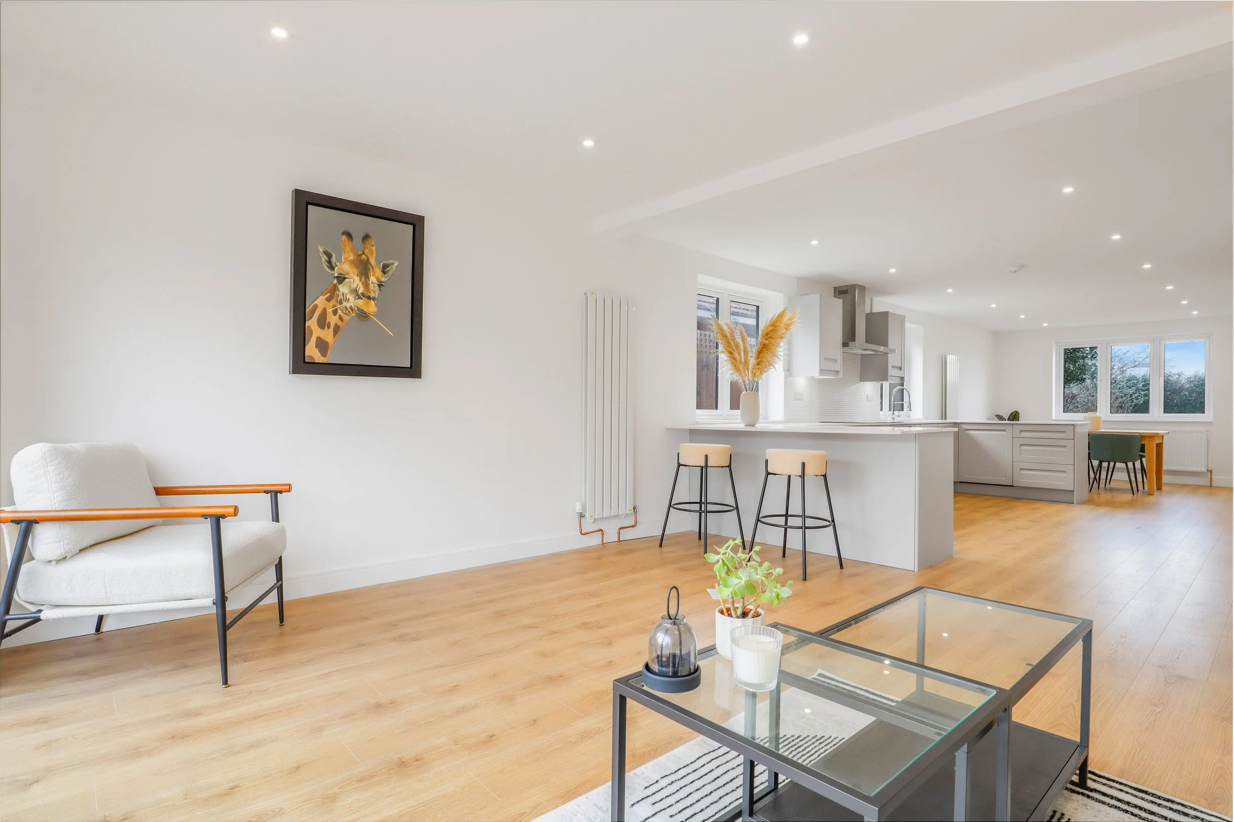 Open-concept living space with white walls, hardwood floors, a white sofa with wooden armrests, a framed giraffe artwork on the wall, a glass coffee table, and a modern kitchen with white cabinetry, a breakfast bar with stools, and a dining area near