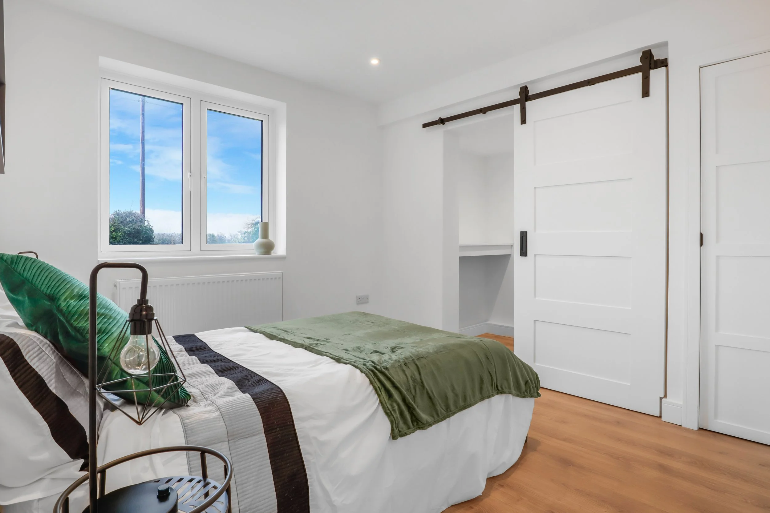 A bedroom with a window, a bed with white, brown, and green bedding, a lamp, and a sliding barn door.