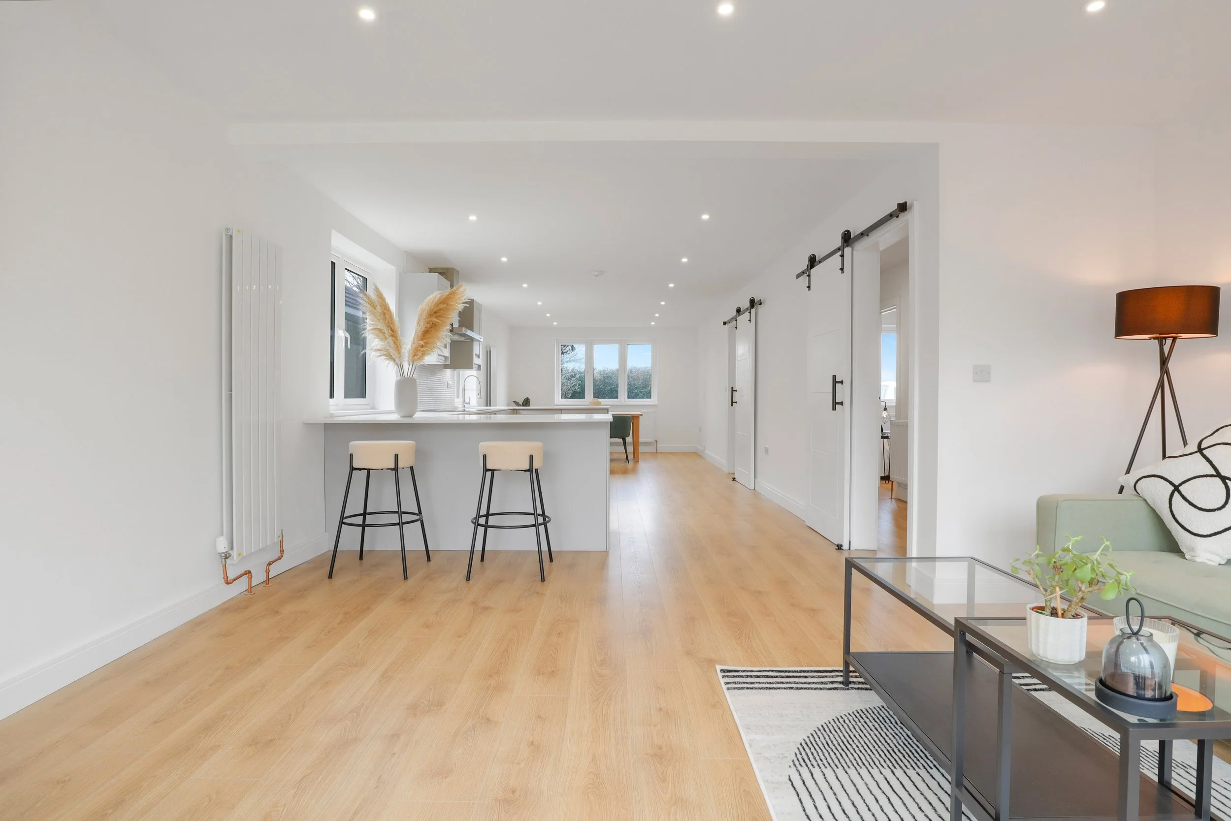 Open-concept living room and kitchen with light wood flooring, white walls, and minimalistic decor. Features a bar area with two beige stools, a white vase with pampas grass, and a black coffee table with decorative items. contains a sofa with black 