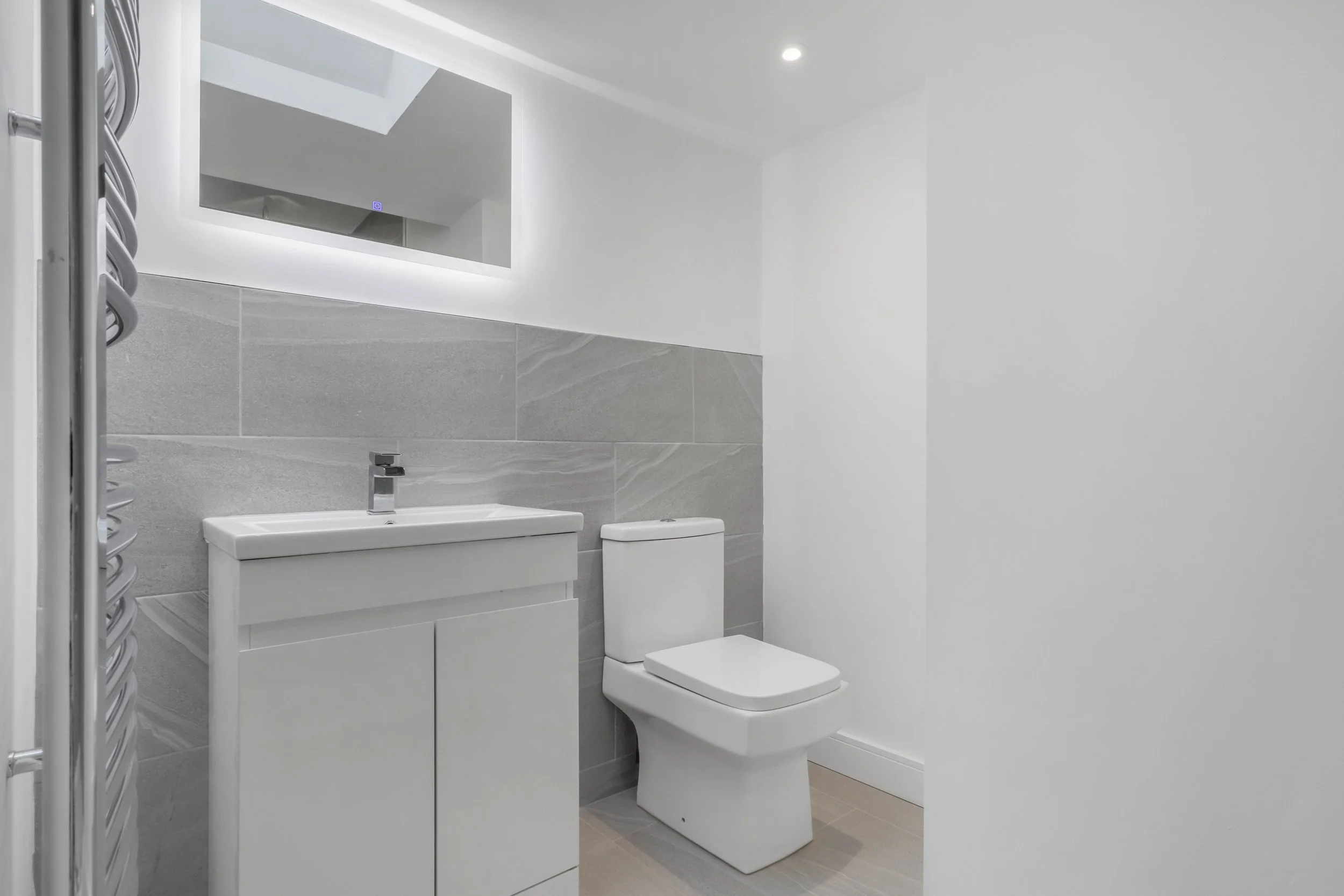 A modern, minimalistic bathroom with a white sink vanity, a toilet, a wall mirror with LED lighting, gray tiled wall, and white-painted walls.