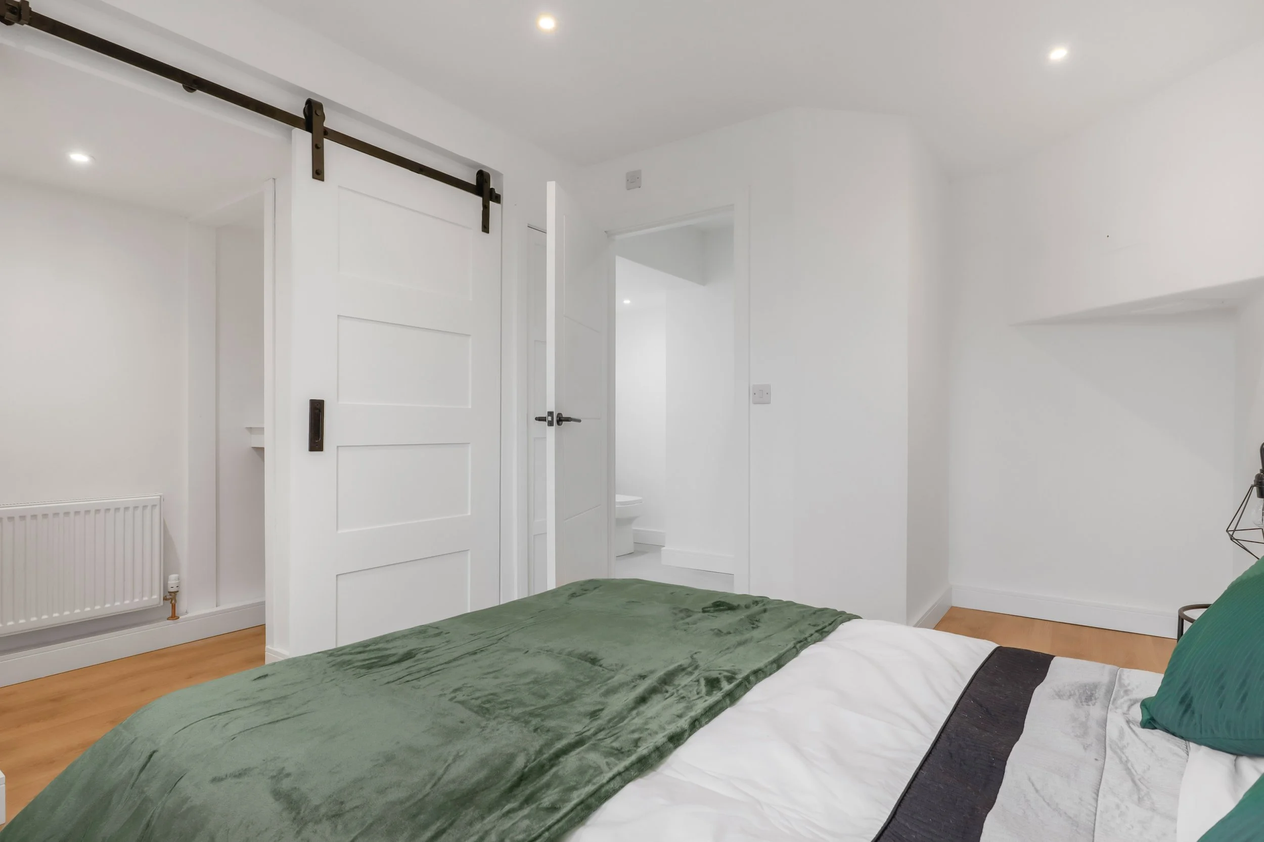 A minimalist bedroom with white walls, a bed covered in a green blanket and pillows, a sliding barn door on a track, and a doorway to a small bathroom with a toilet.