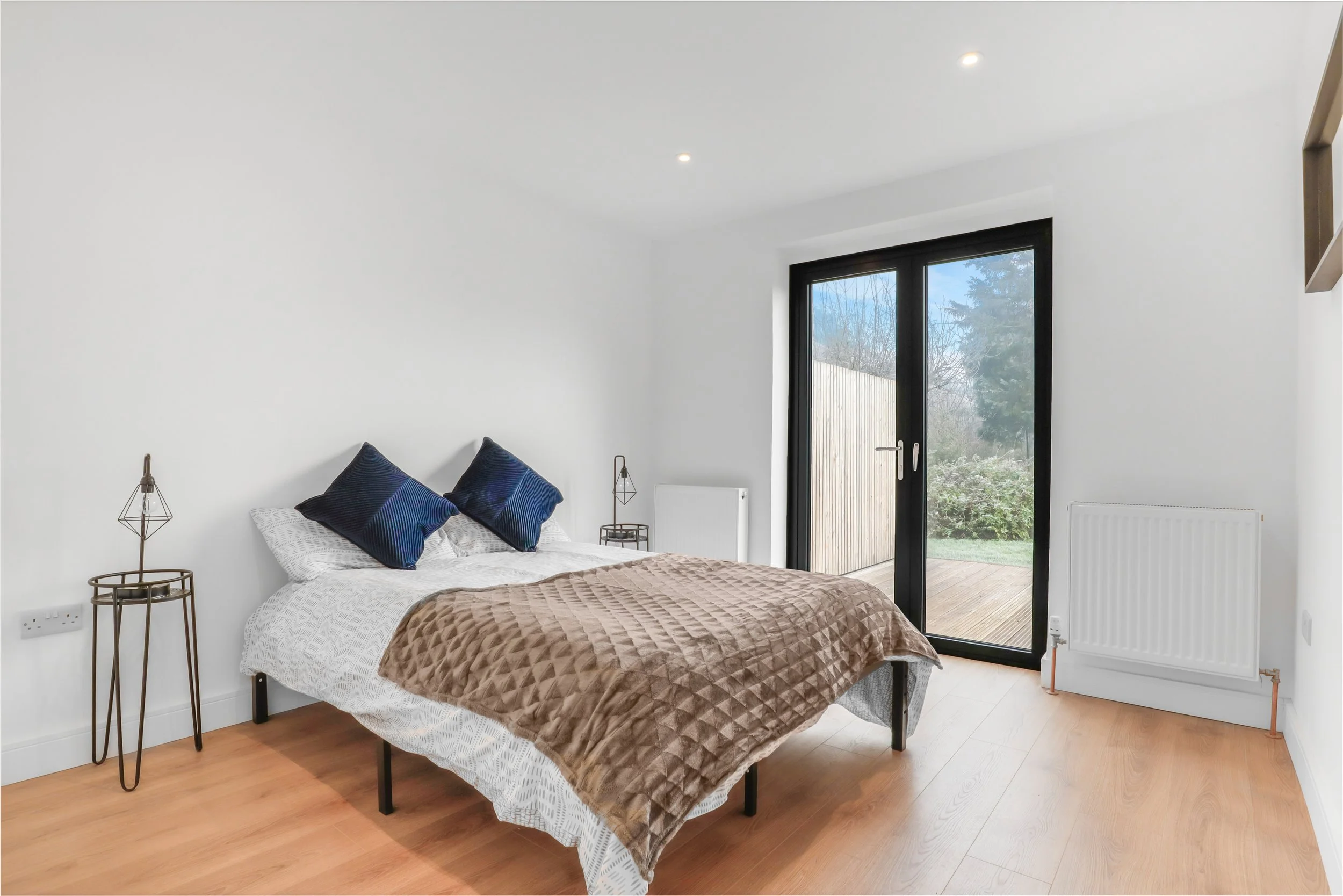 A bedroom with a bed covered in white and brown bedding, two navy pillows, and two side tables with lamps. There is a large glass door leading to an outdoor deck with a garden view.