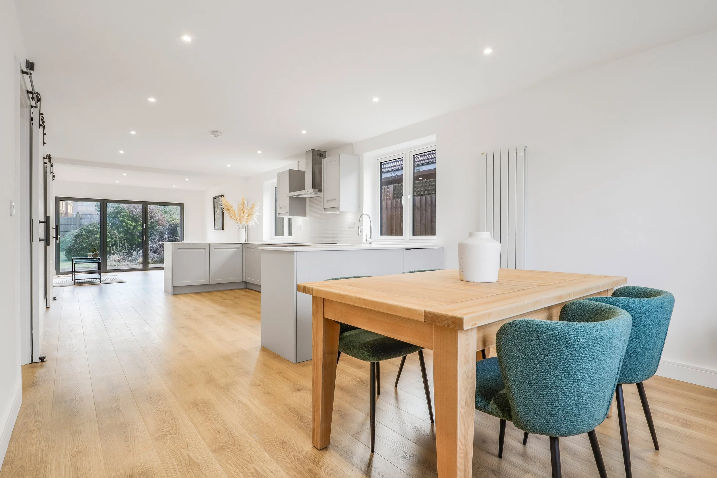 Open-concept kitchen and dining area with wooden floor, white walls, a wooden dining table with four green chairs, a white vase on the table, white kitchen cabinets, a window, and sliding glass doors leading to a backyard with a garden.