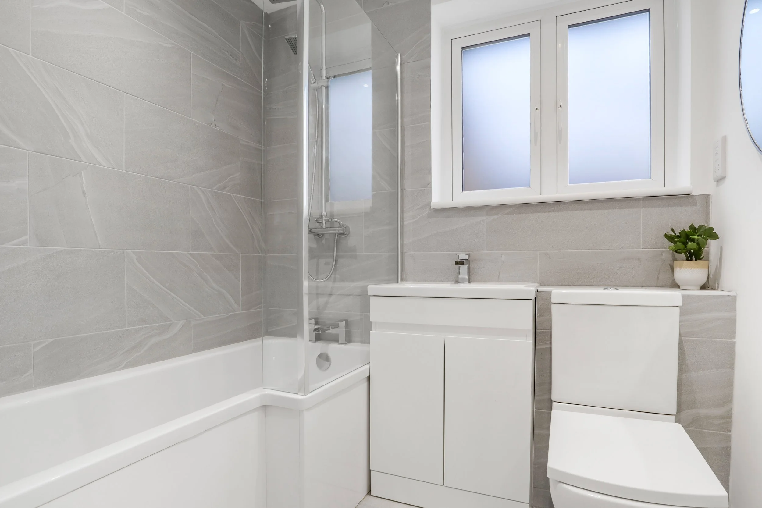 Modern bathroom with a bathtub, a shower with a glass panel, a white vanity with a sink, a toilet, a white window with frosted glass, and a small potted plant on the windowsill.