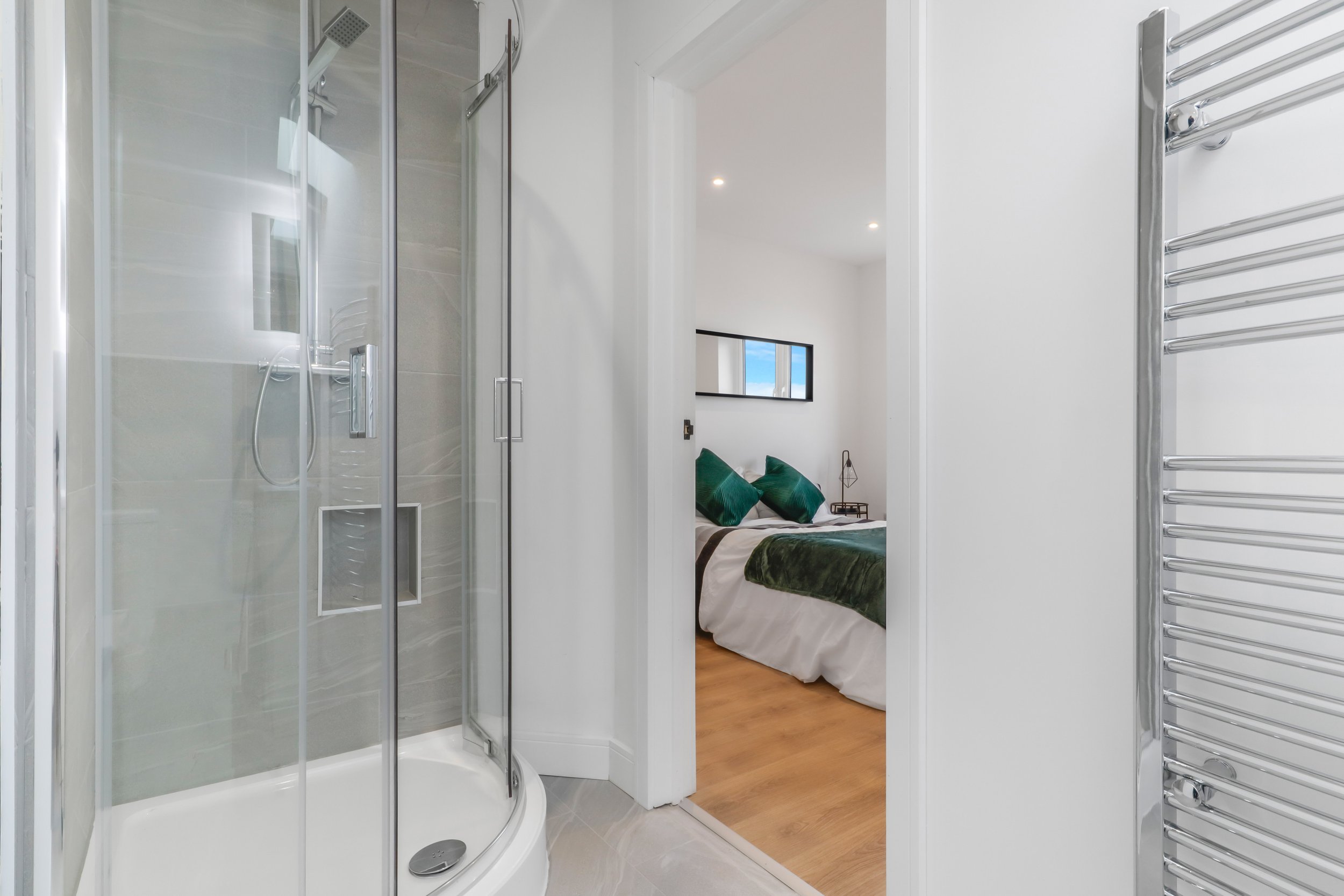 View of a modern bathroom with a glass shower enclosure, white tiled walls, and a heated towel rail on the right, looking into a bedroom with a bed, green pillows, and a small bedside table.