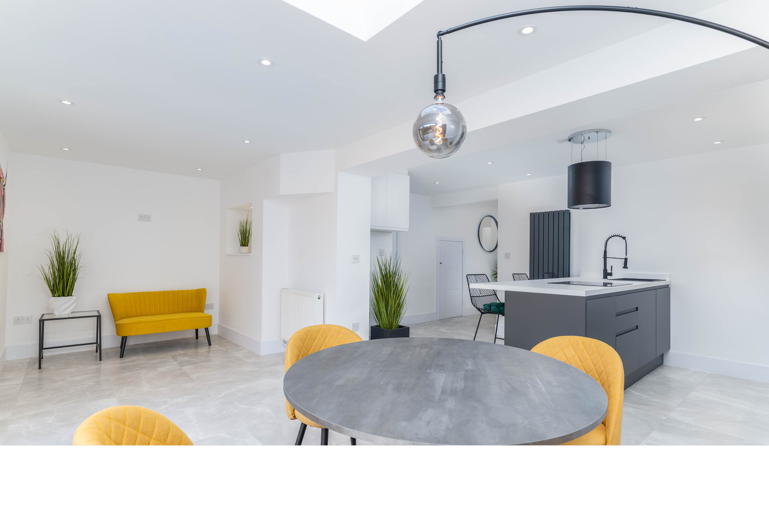 Modern open-concept kitchen and dining area with white walls, a round gray table, mustard yellow chairs, a gray kitchen island, black fixtures, and green plants.