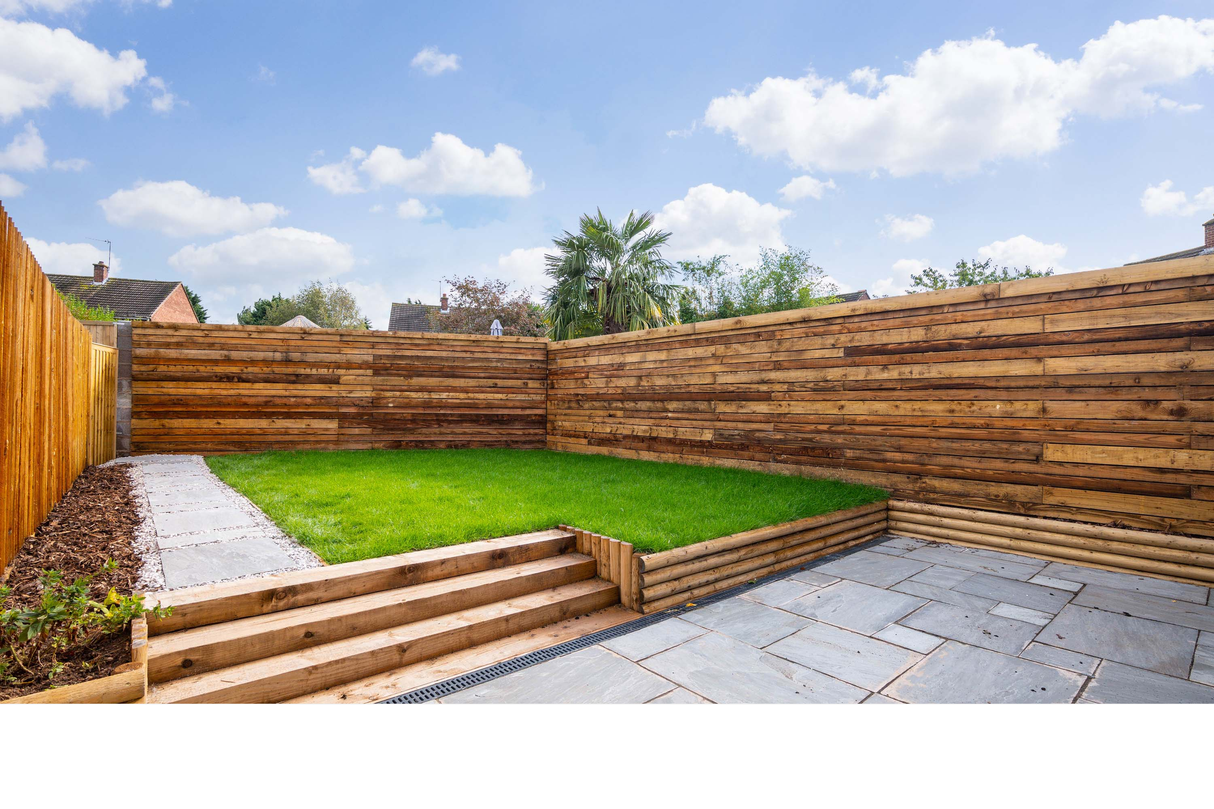 Backyard with a small lawn, stone pathway, wooden steps, and wooden fencing on three sides under a partly cloudy sky.