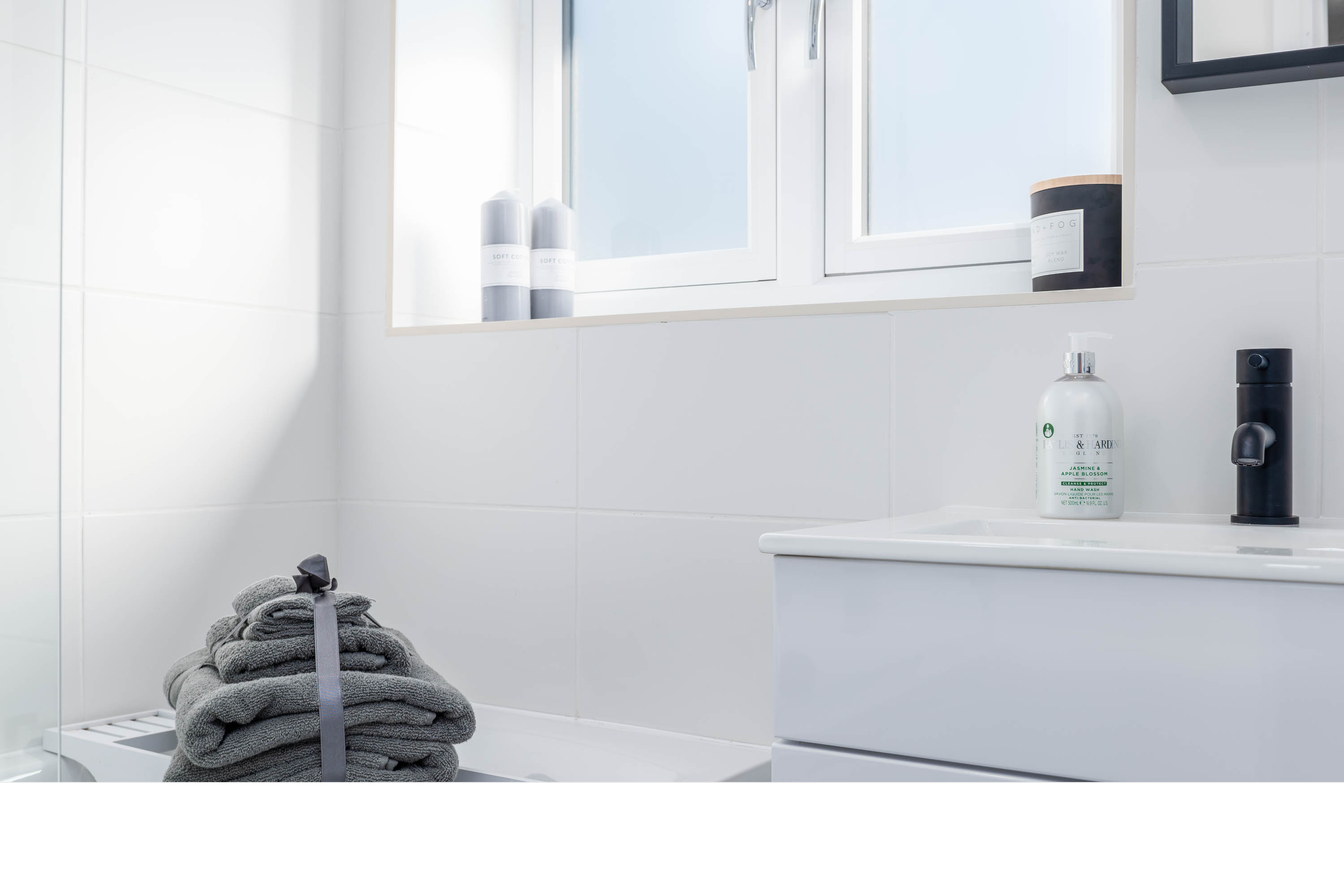A modern bathroom with white tiles, a window allowing natural light, a stack of gray towels tied with a ribbon on a heating radiator, a sink with a black faucet, and various bottles of toiletries on the windowsill and sink.