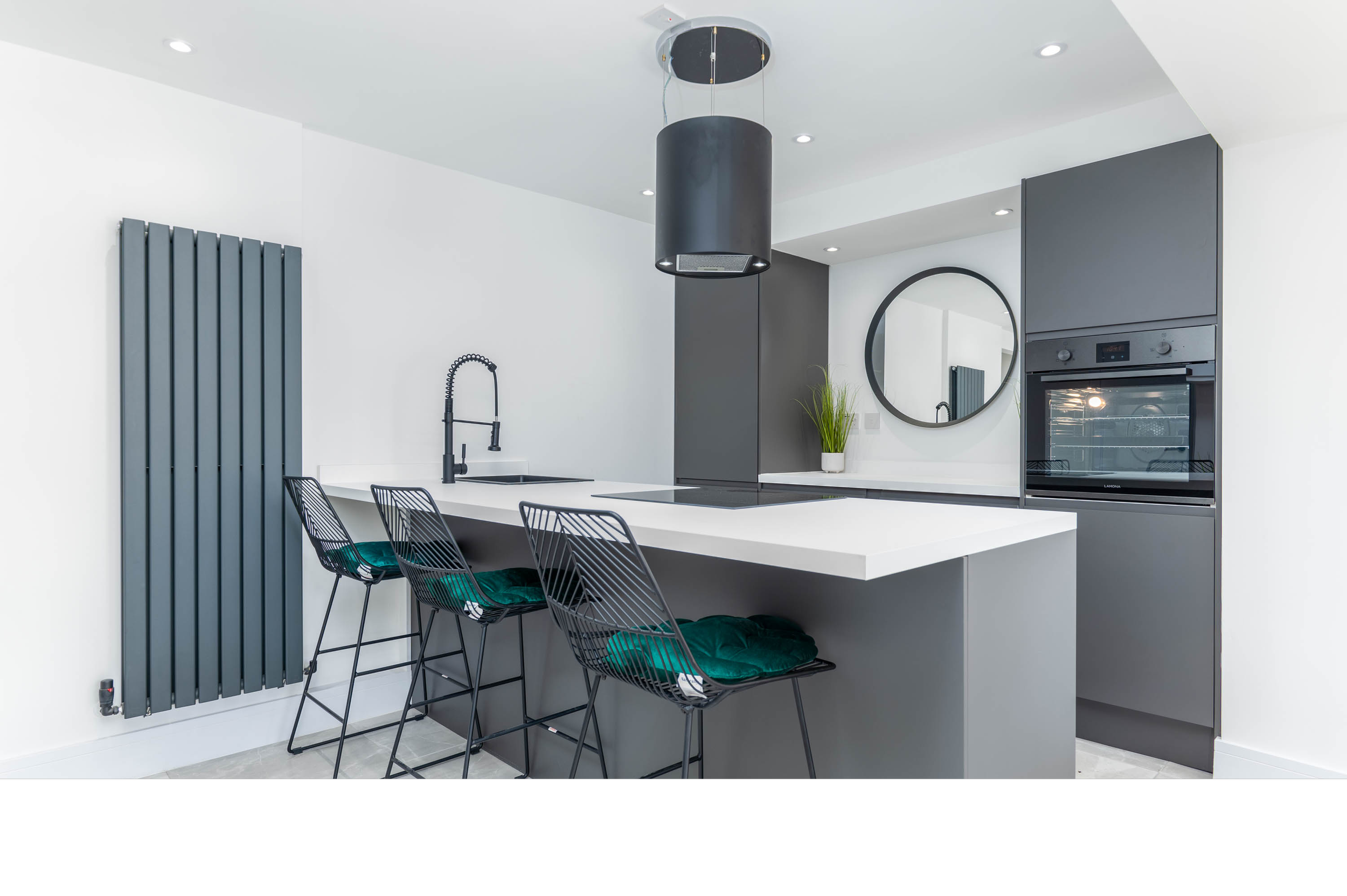 Modern kitchen with black and white color scheme, featuring a white island with black bar stools with green cushions, black cabinets, built-in oven, round wall mirror, and a black cylindrical pendant light hanging from the ceiling.