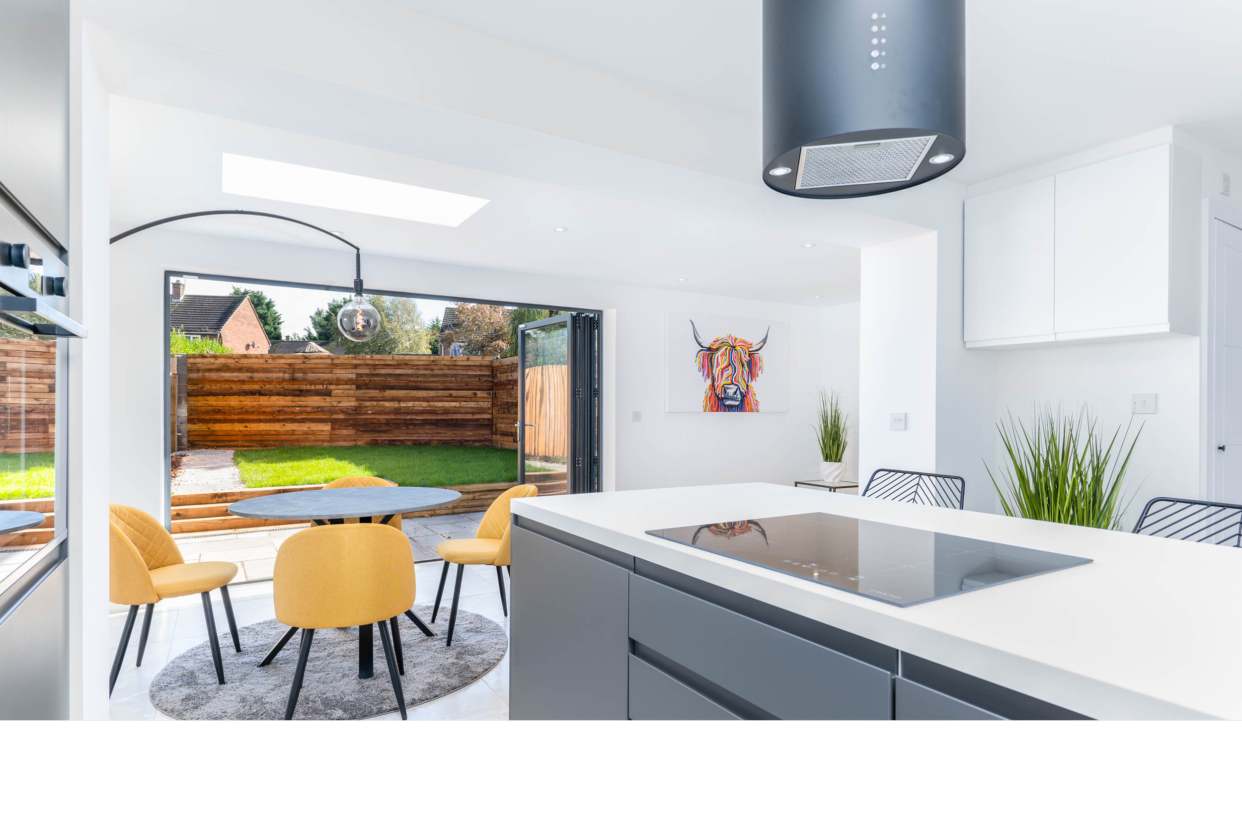 Modern open-concept kitchen with white cabinetry, a large kitchen island with a built-in stove, black chairs, a dining area with yellow chairs and a round table, and large sliding glass doors leading to a backyard with a wooden fence and green grass.