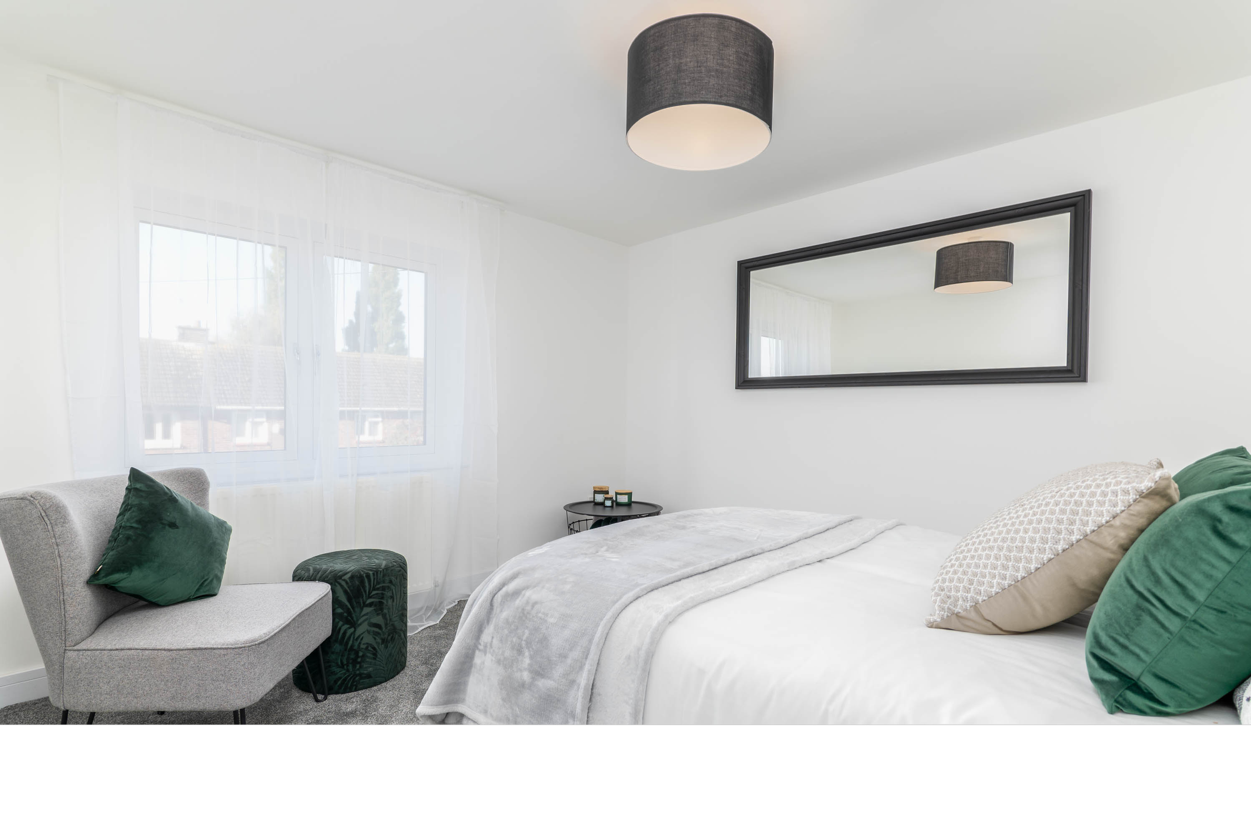A bright bedroom with white walls, a large bed covered in white bedding and gray blankets, green and patterned pillows. There is a window with sheer curtains, a black ceiling light, a black-framed mirror on the wall, a gray chair with pillows, and a 
